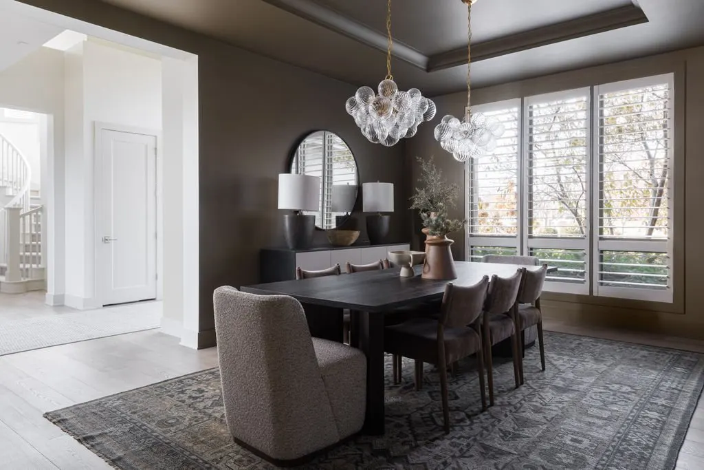 Modern dining room with a dark wood table, eight chairs, a large vase with greenery, and a rug. Bright light from large windows with white shutters, white walls, and decorative light fixtures hanging from the ceiling.