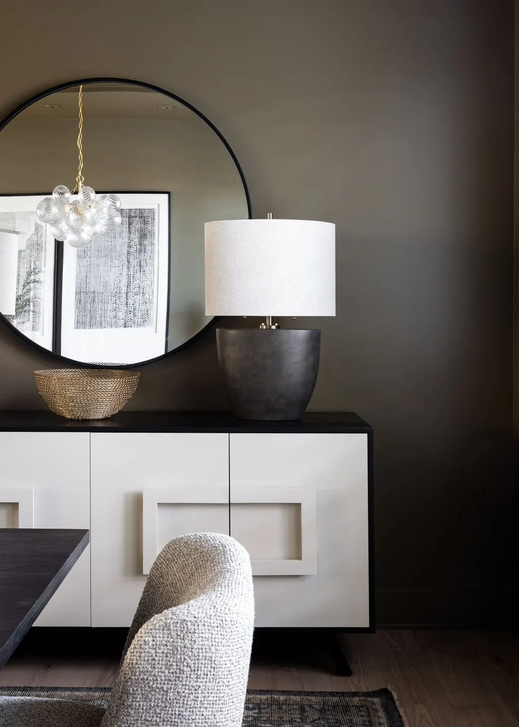 A modern sideboard with a circular mirror, a table lamp, a woven bowl, and a seating chair in a contemporary living room.