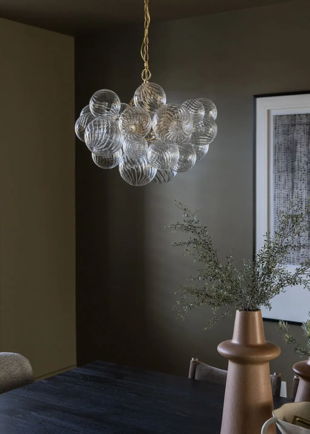 A modern chandelier made of clear glass globes hanging above a dark wooden dining table. A beige vase with green foliage is on the table, and framed art is on the wall in the background.