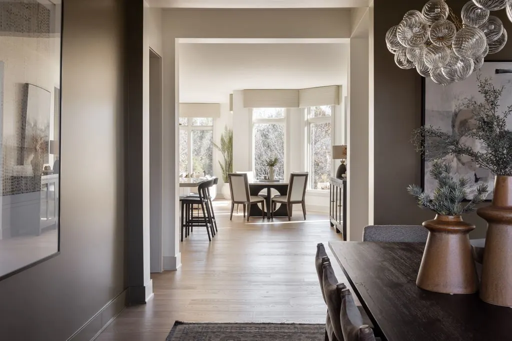 View of a dining area in a home with large windows, a round dining table, and chairs, illuminated by natural light.