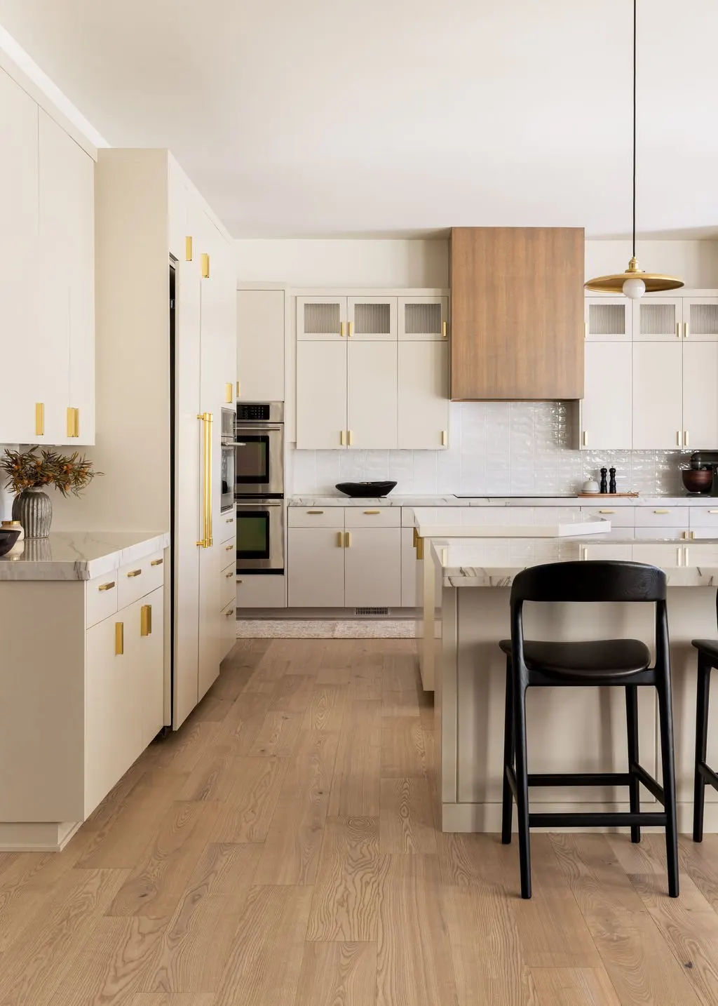 Modern kitchen with white cabinets, gold handles, a wooden range hood, black chairs, and light wood flooring.