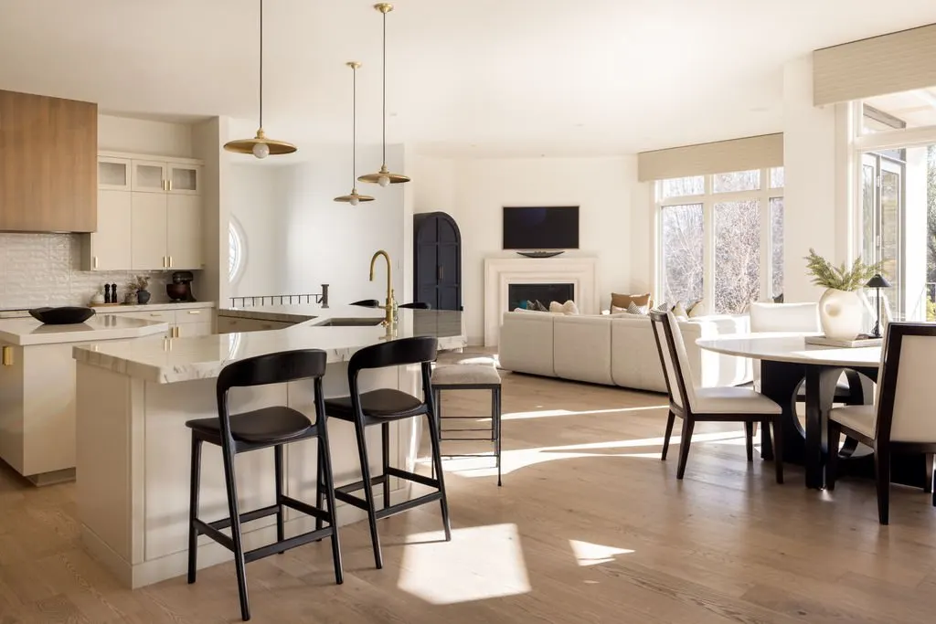 Open-concept kitchen and living room with modern decor, white cabinets, black chairs, and large windows letting in natural light.