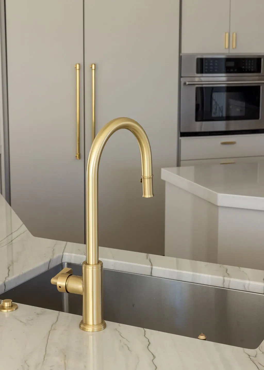 A modern kitchen with a black sink, gold faucet, white marble countertop, gray cabinets with gold handles, and stainless steel built-in oven.
