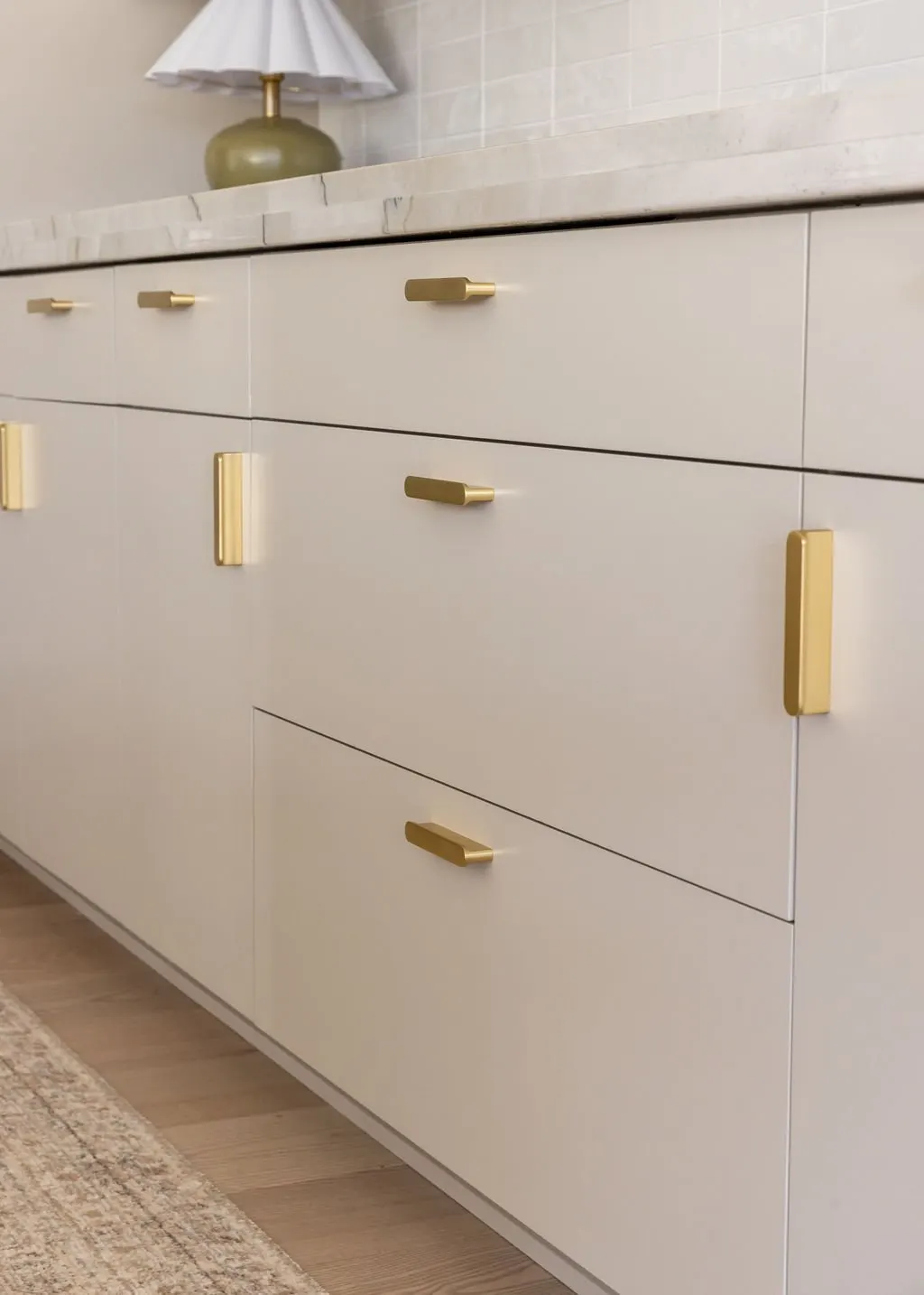 Close-up of a modern white cabinet with gold handles, atop a marble countertop. A green ceramic lamp with a white shade is on the counter against a tiled wall.