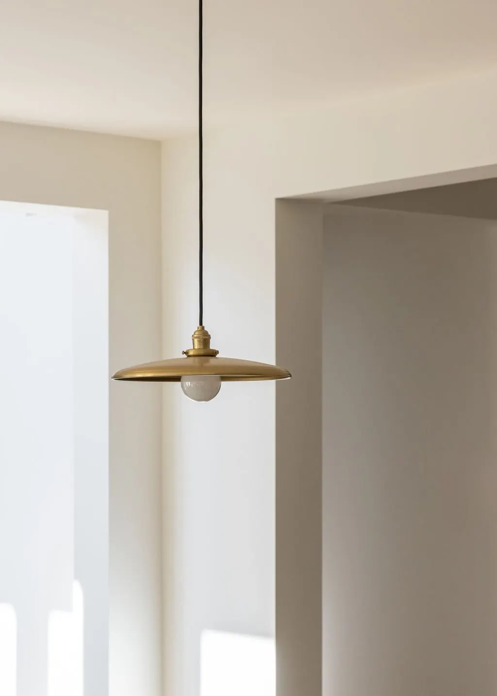 Close-up of a modern ceiling light fixture with a brass finish hanging from a black cord in a minimalist room with white walls.