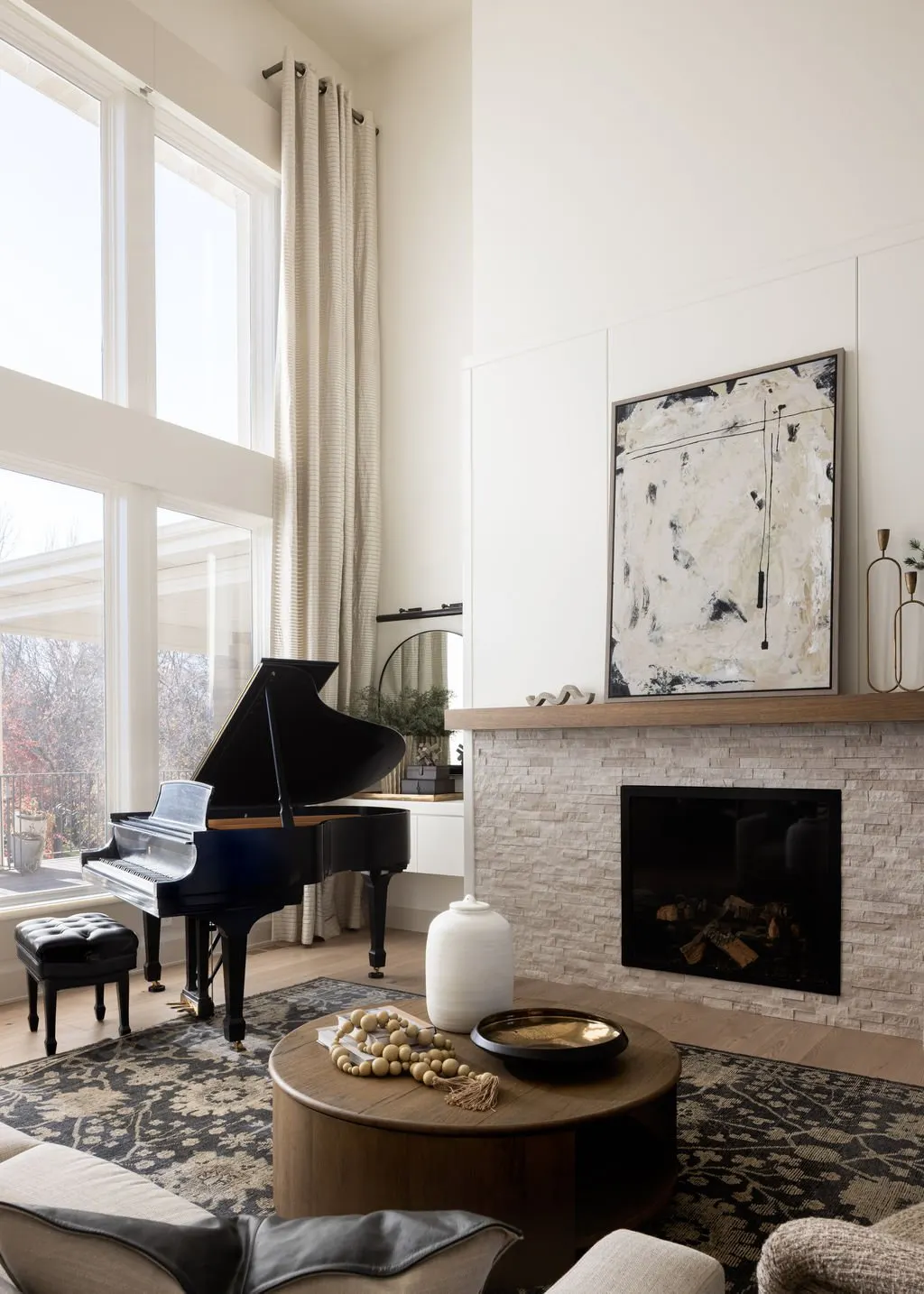 Living room with large windows, white curtains, black grand piano, abstract artwork above fireplace, and neutral-toned decor.