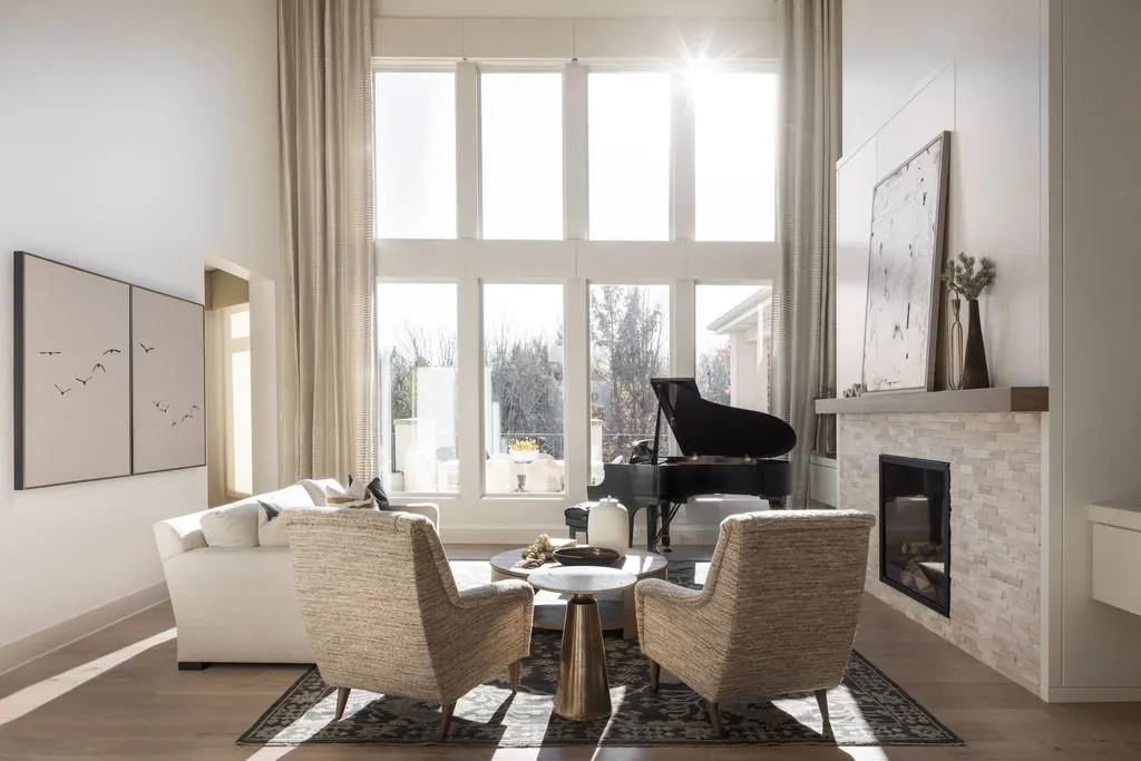 Living room with large windows, white sofa, two beige armchairs, black grand piano, fireplace, artwork, and decorative vases, bright sunlight streaming in.