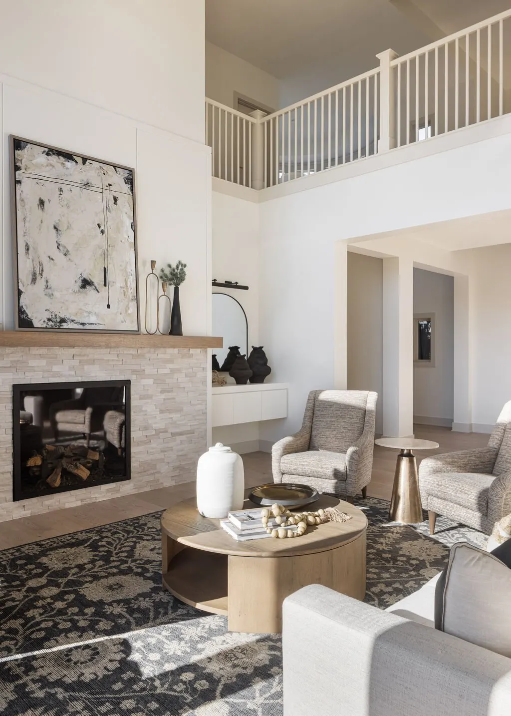 Modern living room with a fireplace, abstract artwork above it, neutral-colored armchairs, round coffee table with decorative items, area rug, and a second-floor balcony with white railing.