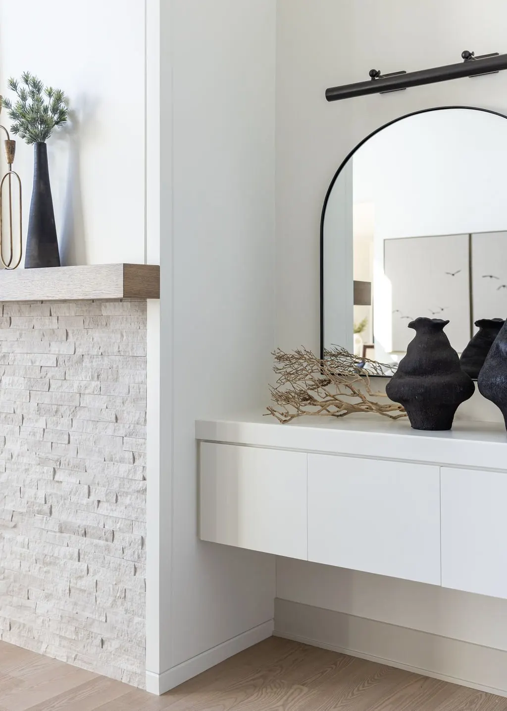 Decorative vases and driftwood on a white floating cabinet in front of a rounded mirror in a modern, minimalist interior.