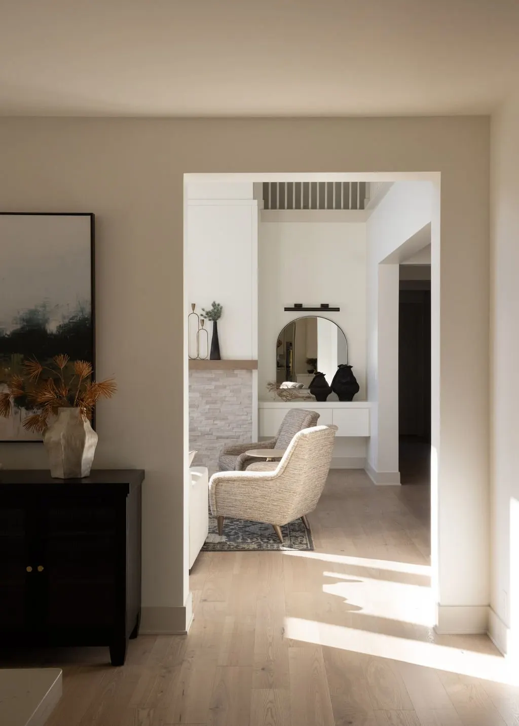 Living room view through a doorway showing beige armchair, decorative vases and mirror, with sunlight on wooden floor.