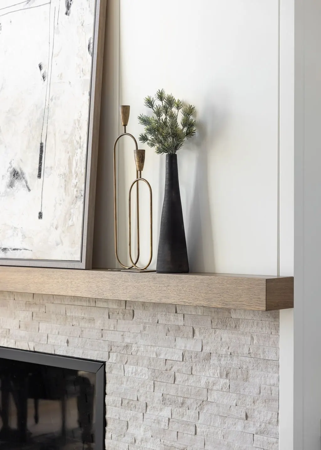 A decorated modern interior with a wood mantel, a black tall vase with greenery, a gold abstract sculpture, a framed abstract painting, and a stone fireplace.