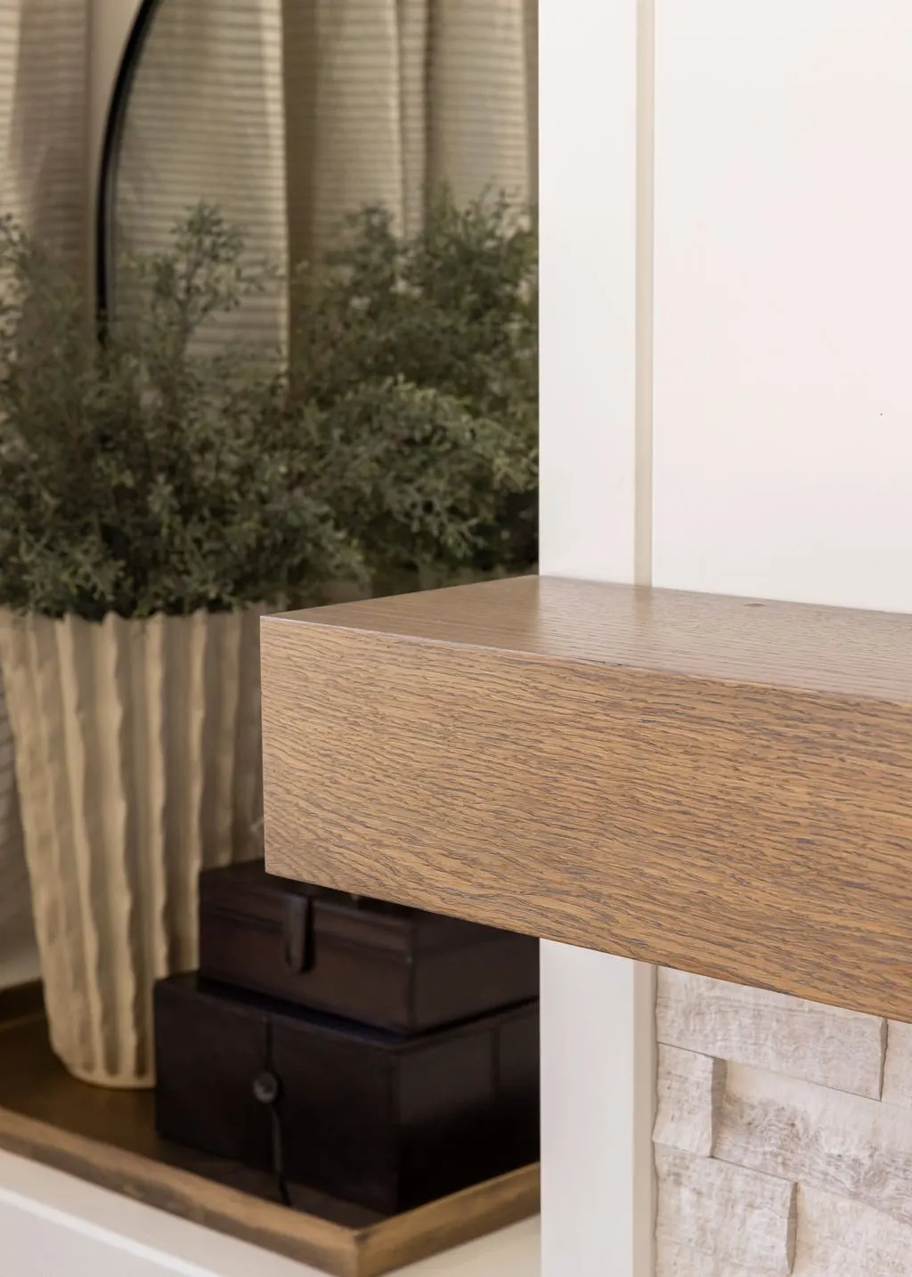 Close-up of a wooden shelf next to a white wall in a room with a large window and a potted plant.