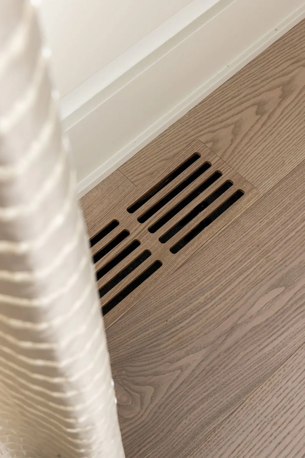 Close-up of floor vent cover with wooden slats on hardwood floor near white baseboard and wall.