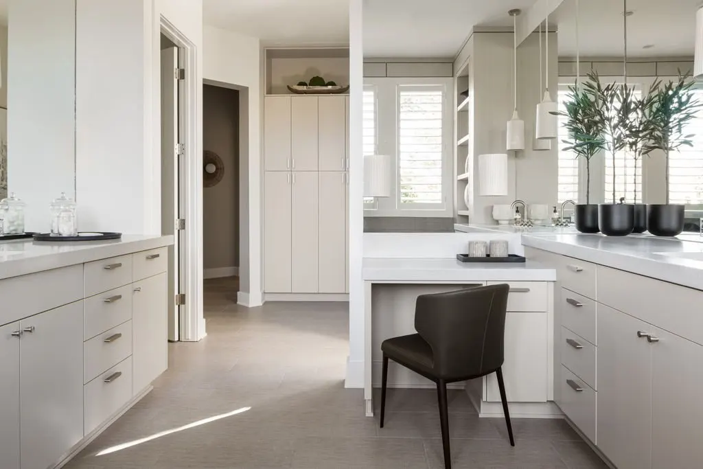 Modern kitchen with white cabinets, a white countertop, pendant lights, and a large window with white shutters. There are black planters with greenery on the right and some glass jars on the left. A black chair is placed at a small desk area.