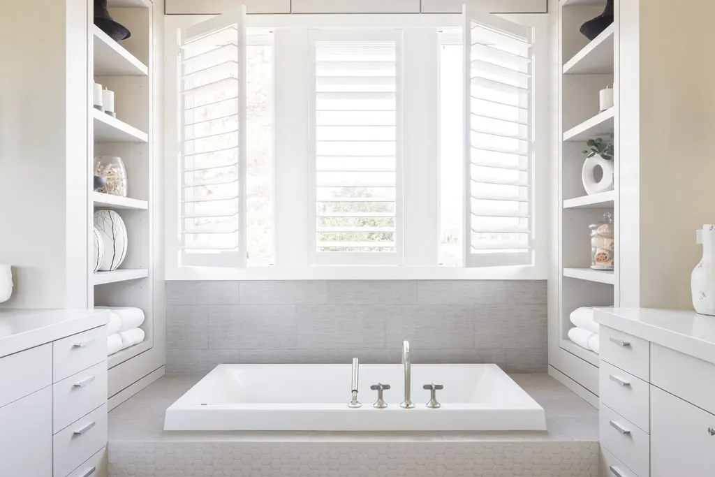 Bright bathroom with a built-in bathtub, surrounded by white cabinetry and shelves filled with towels and decorative items, large windows with plantation shutters allowing natural light.