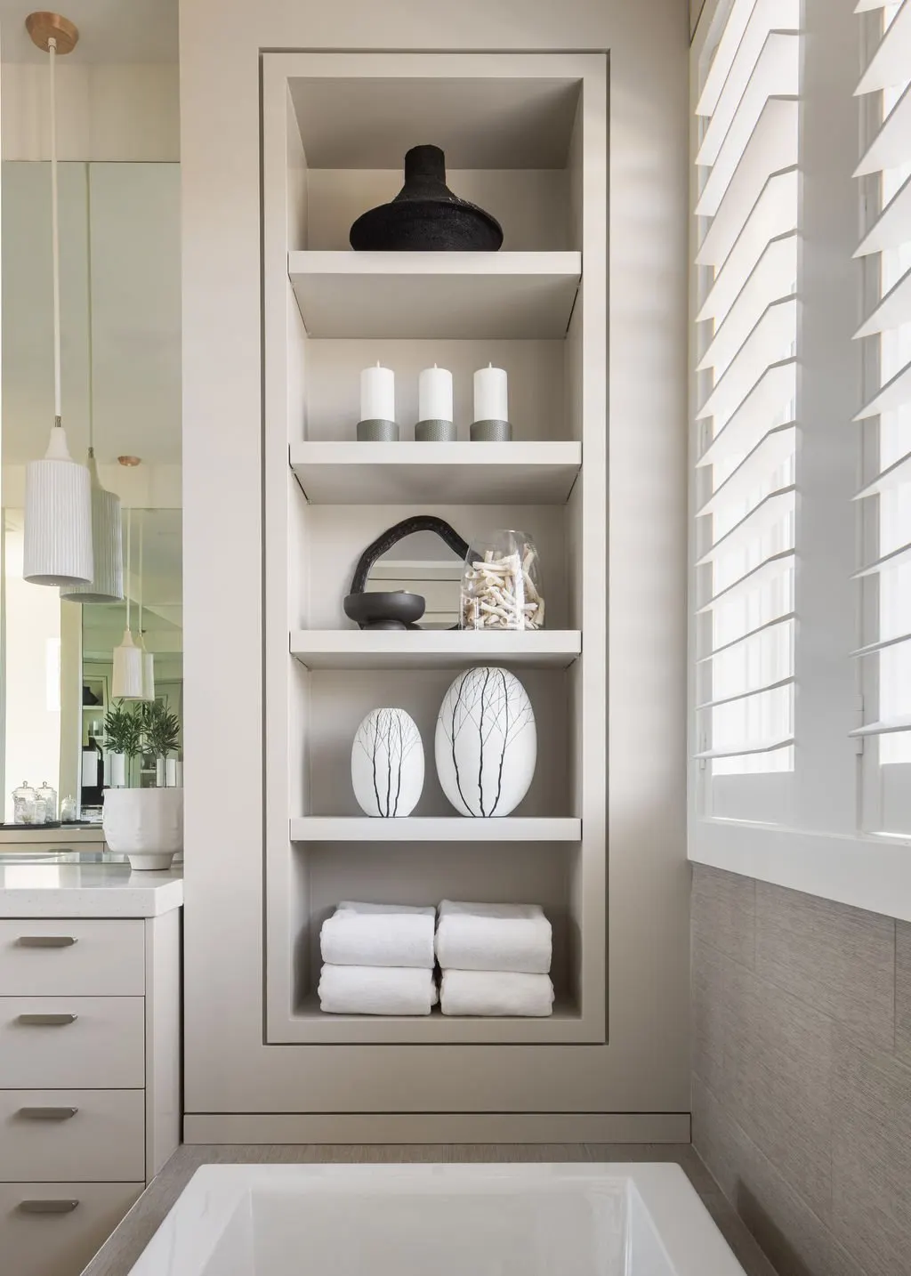 Built-in white shelving unit with decorative objects and towels in a bathroom, near a window with white plantation shutters.