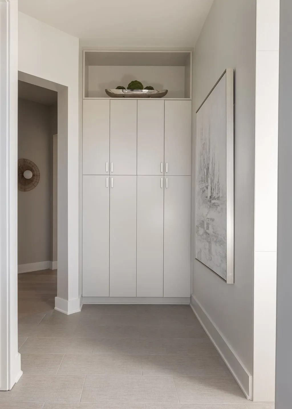 White built-in cabinet against a wall with a decorative shelf on top holding broccoli and a decorative bowl. A large abstract painting is on the right wall.