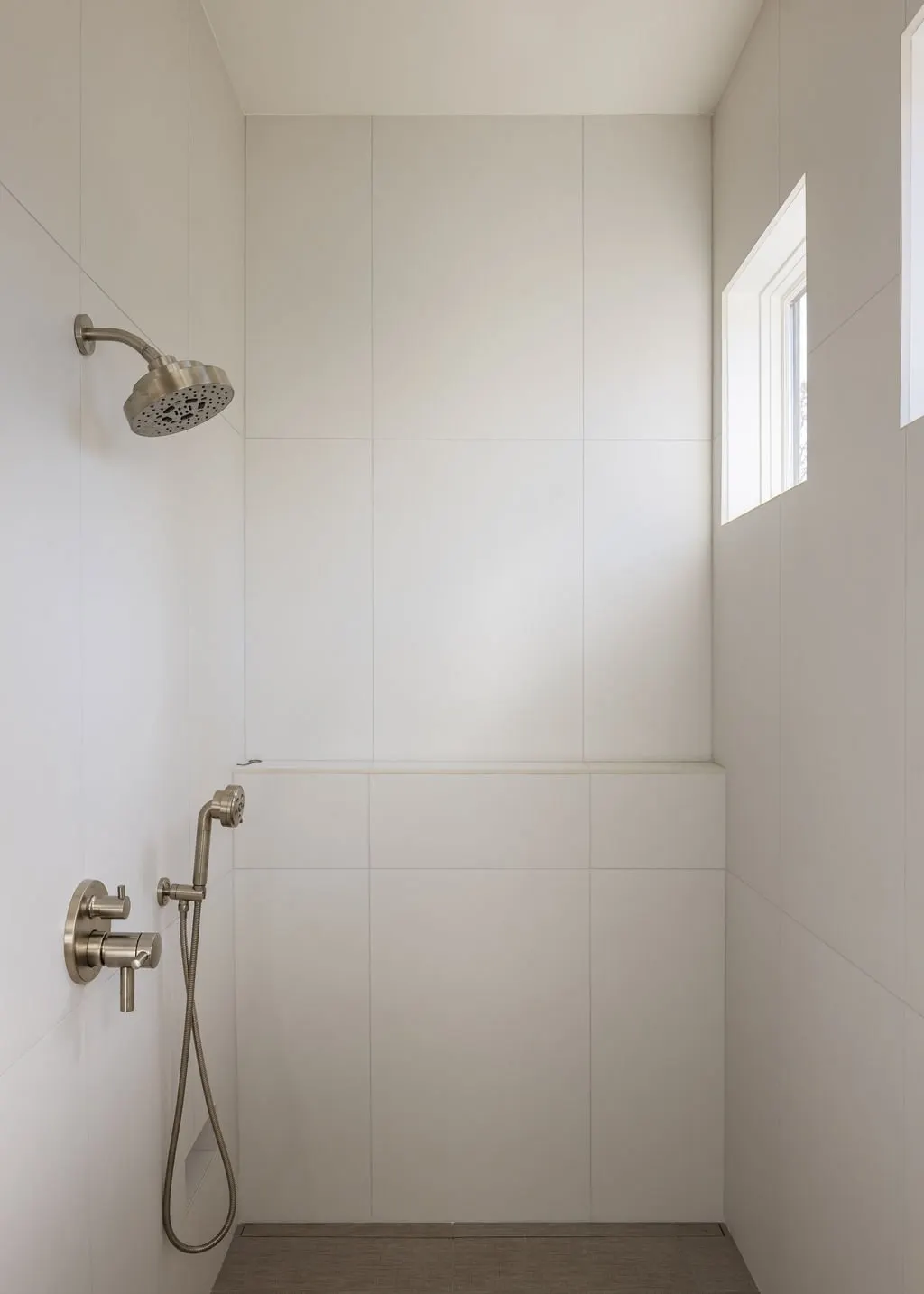 Empty shower with a wall-mounted showerhead, a handheld shower attachment, and a small window in a modern, minimalist bathroom.