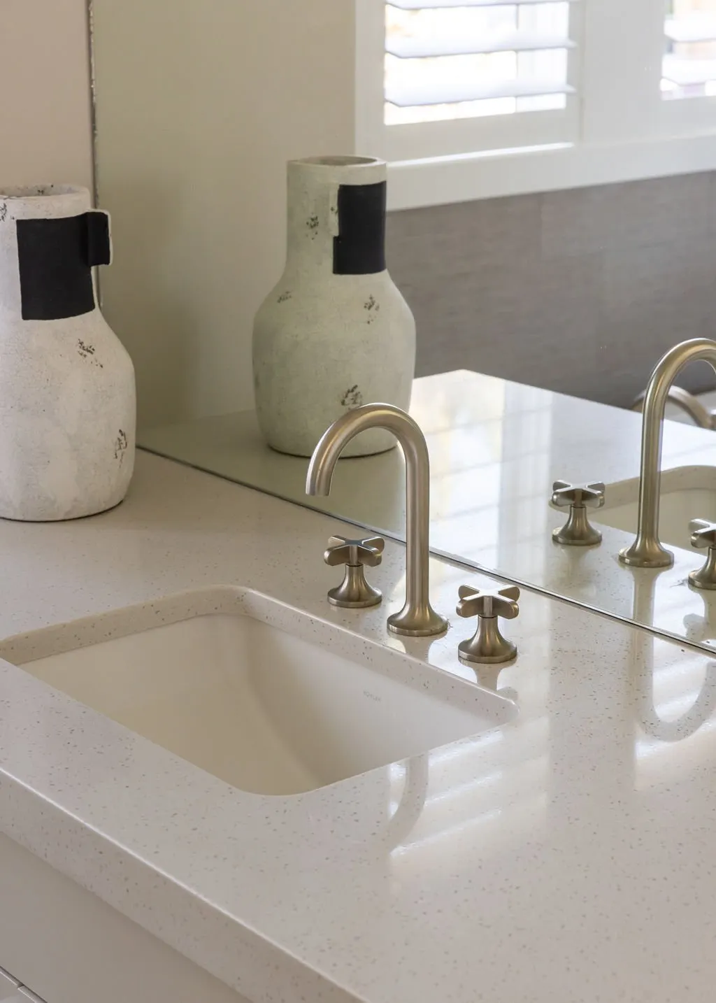 A bathroom countertop with a built-in sink, a mirror, two decorative vases, and a window with white shutters in the background.