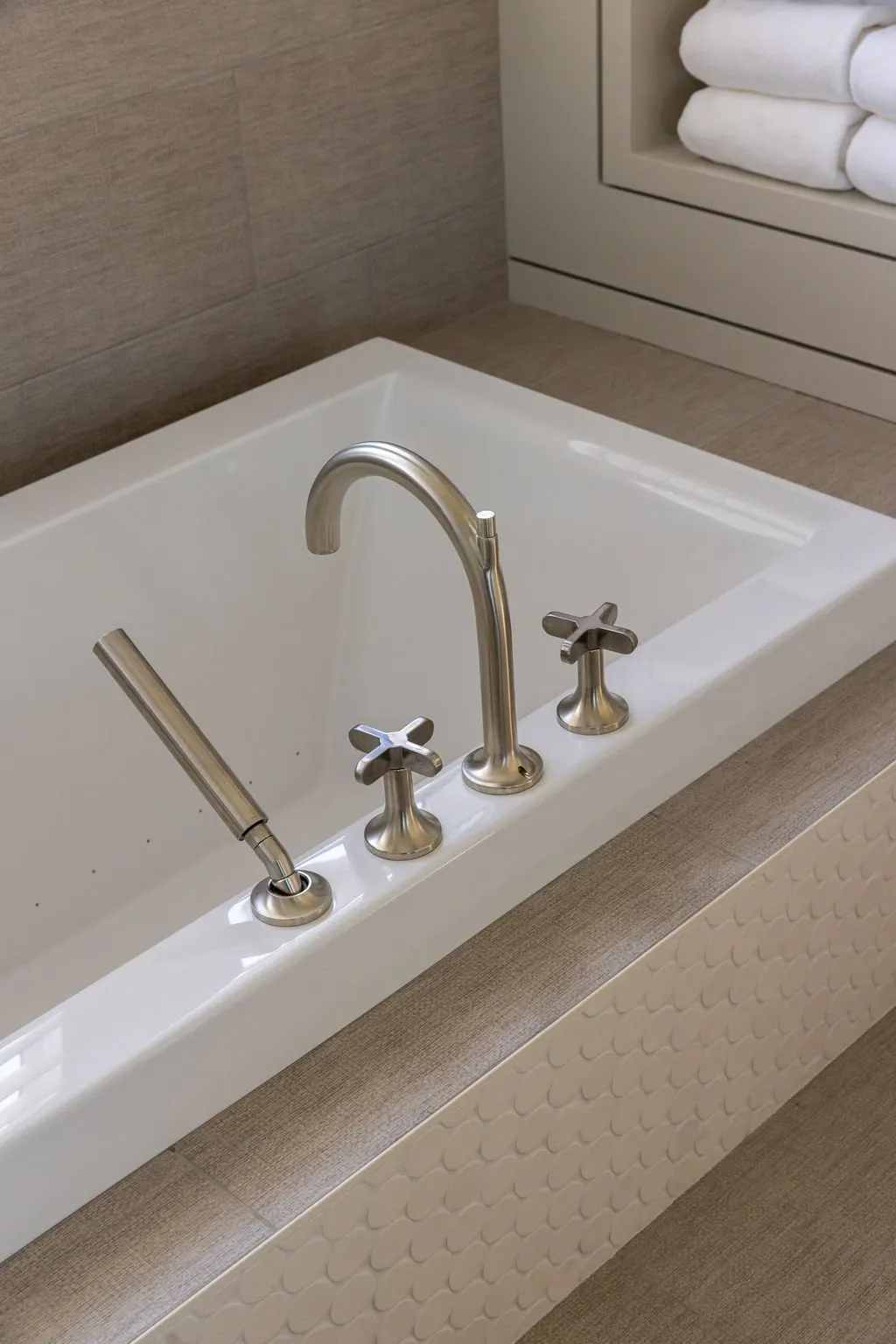 A white bathtub with silver faucet and handles, located in a bathroom with beige tiled walls and countertop, with a shelf holding folded white towels.
