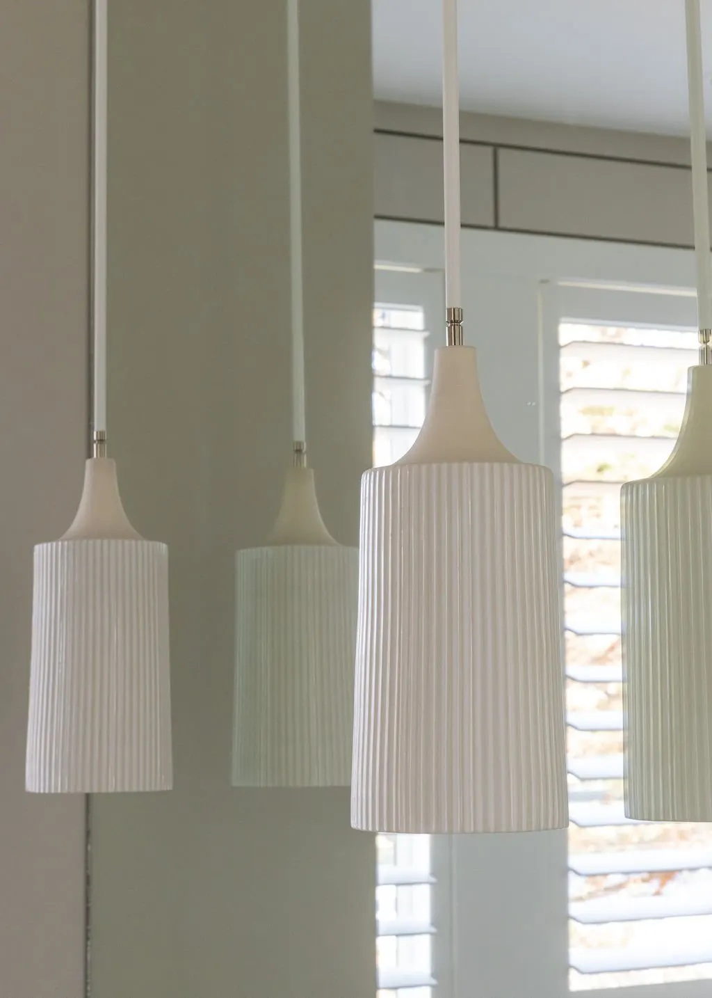Three modern white pendant light fixtures hanging in front of a mirror, with window blinds visible in the background.