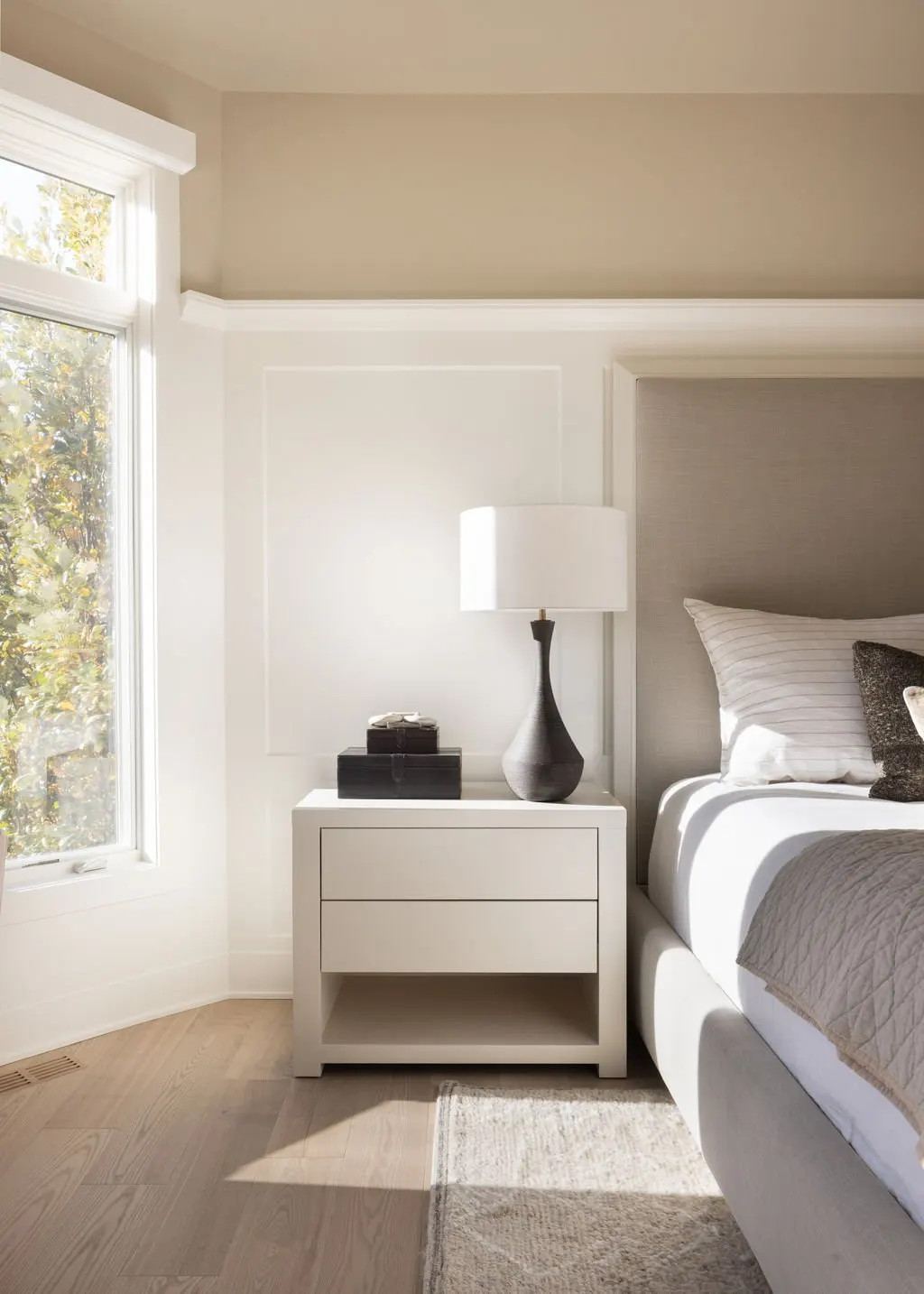 A bedroom with a window showing autumn leaves, a white nightstand with a black box and a gray lamp, a bed with white and gray bedding, and a beige area rug.