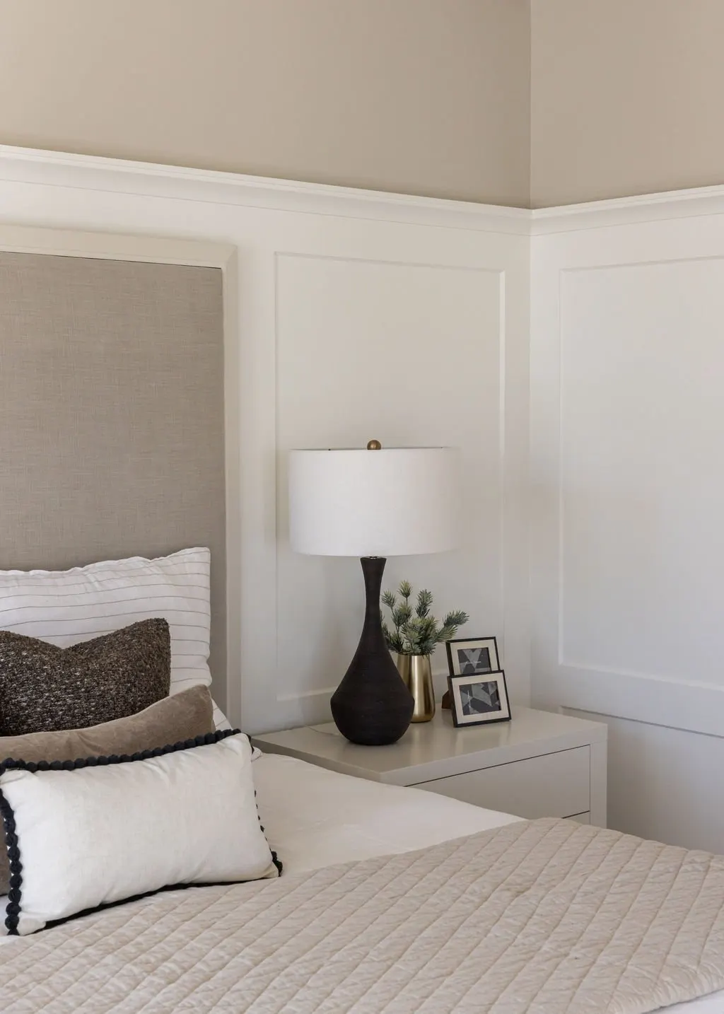 Close-up of a bedroom corner with white bed, pillows, beige quilt, bedside table, black table lamp with white shade, small plant, and picture frames.
