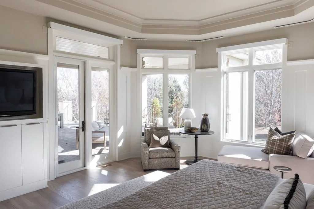 A bright bedroom with large windows, a bed, a small chair, a side table with a lamp, and a sitting area with cushions, overlooking a wooden deck outside.