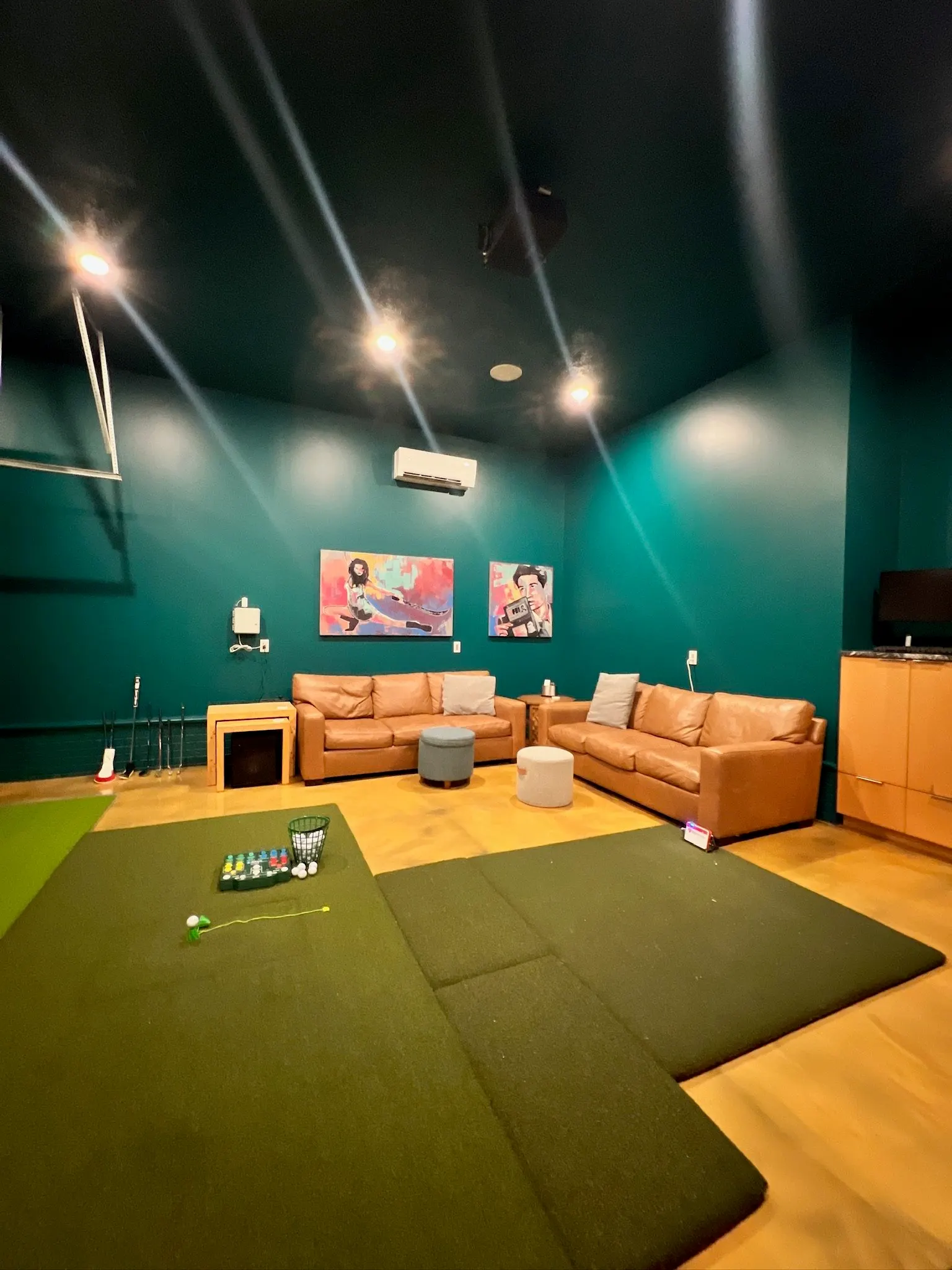 Indoor recreation room with green walls, ceiling spotlights, a brown leather sectional sofa, two small round ottomans, wall art featuring a woman and a man, a golf putting green, and a wall-mounted air conditioner.