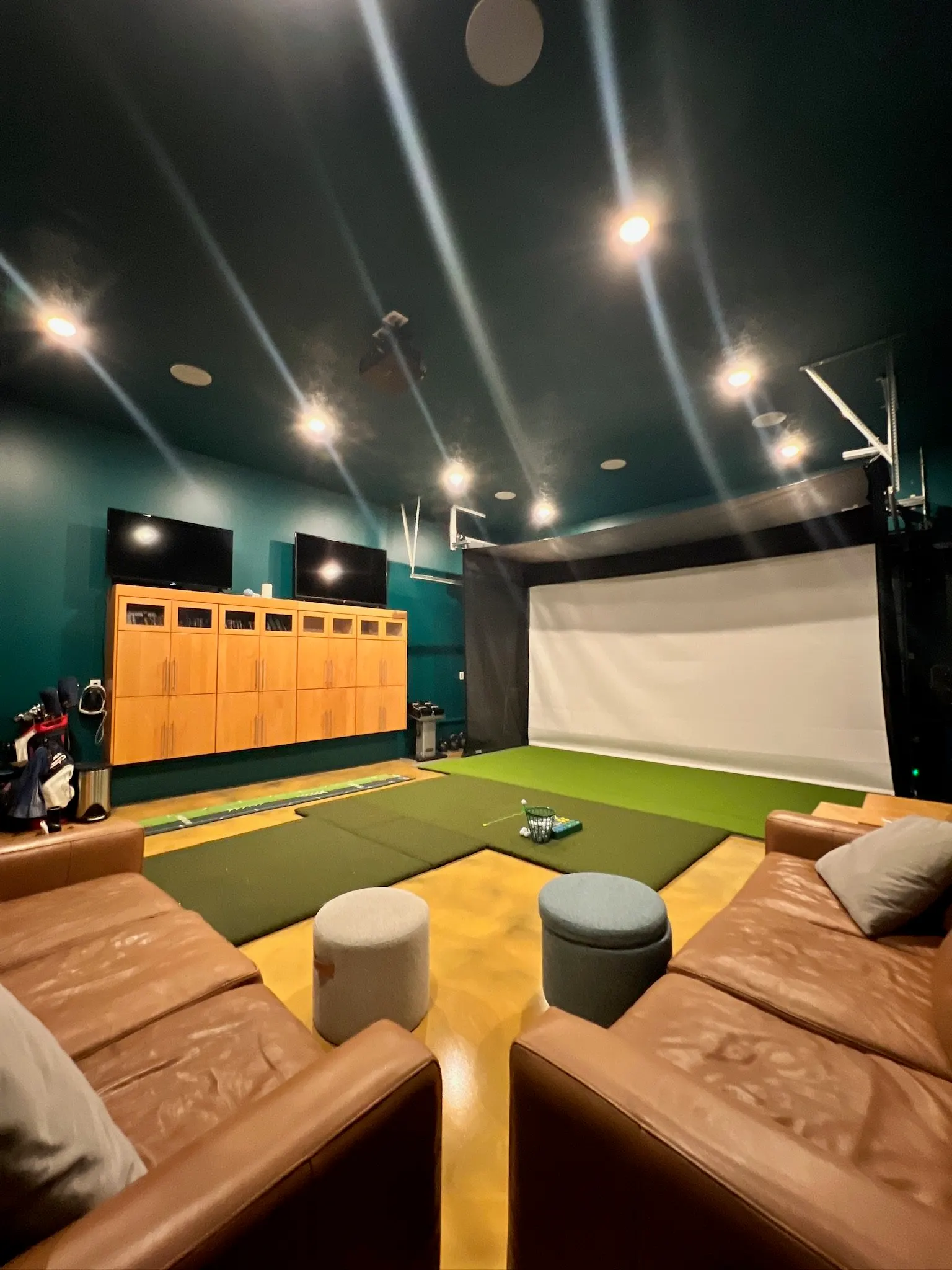Indoor golf simulation room with two leather couches, small round ottomans, a hitting area with a golf mat, a large screen, and multiple monitors.
