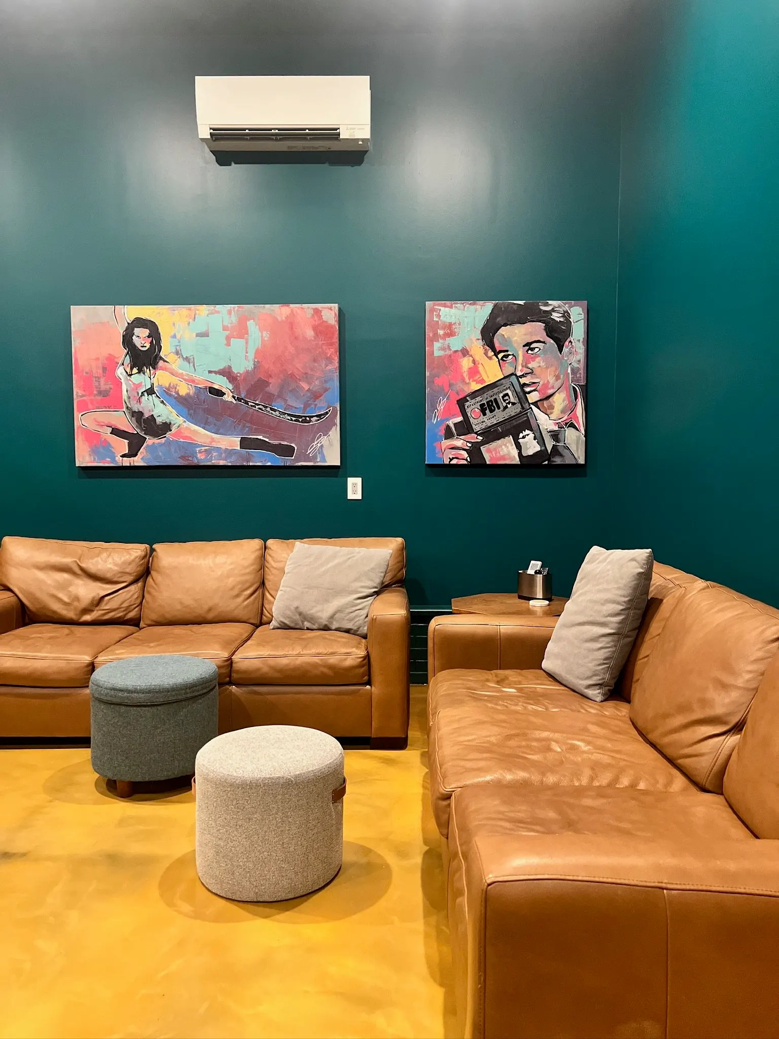 A cozy living room with teal walls, two brown leather sofas with beige pillows, two round ottomans in gray and beige, wall art featuring colorful pop art paintings of a woman with a whip and a man holding a camera, an air conditioner mounted high on
