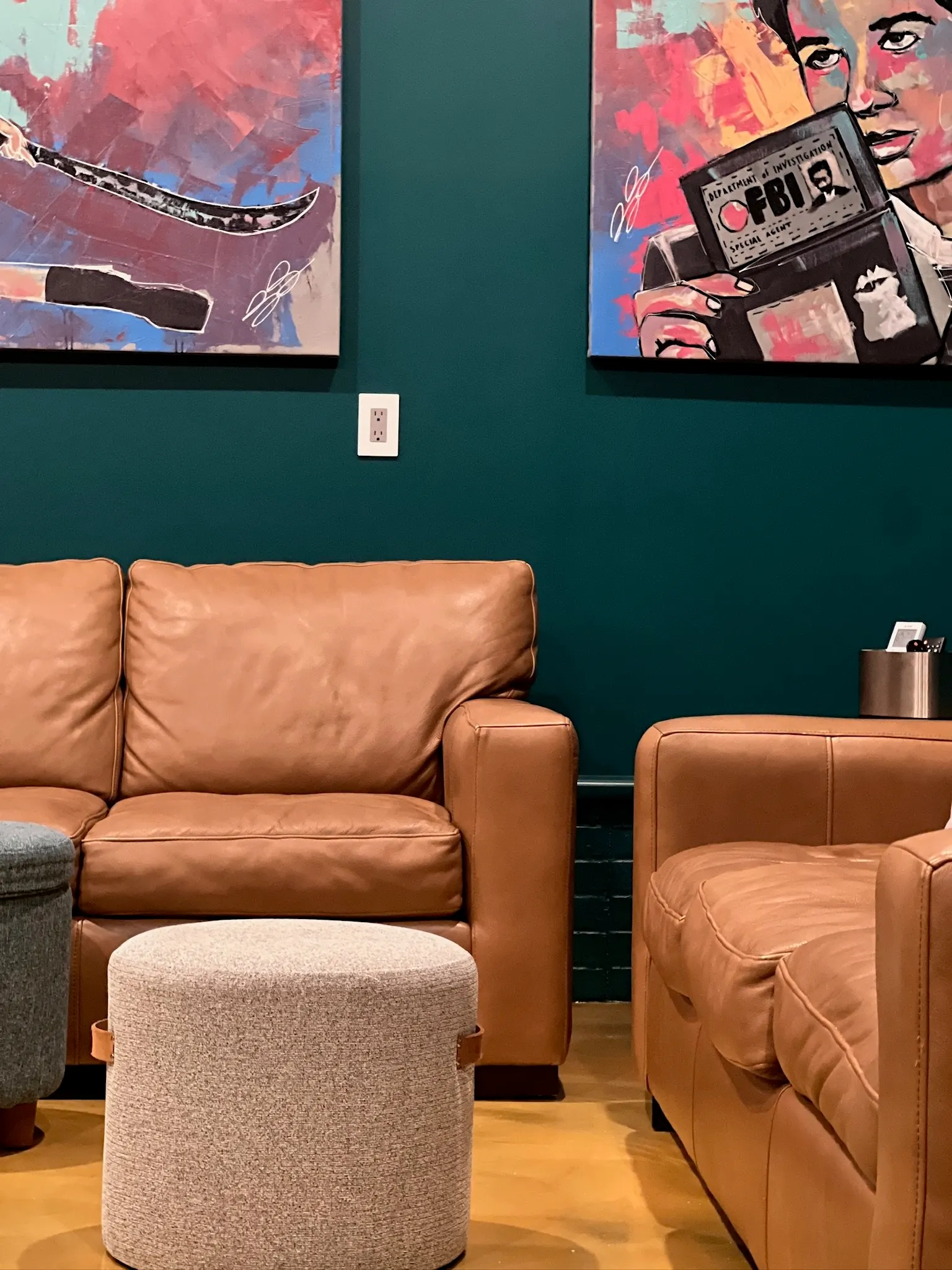Living room with tan leather couch, a matching armchair, round beige ottoman, teal wall with two colorful abstract paintings, and a small side table with a black holder and tissue box.