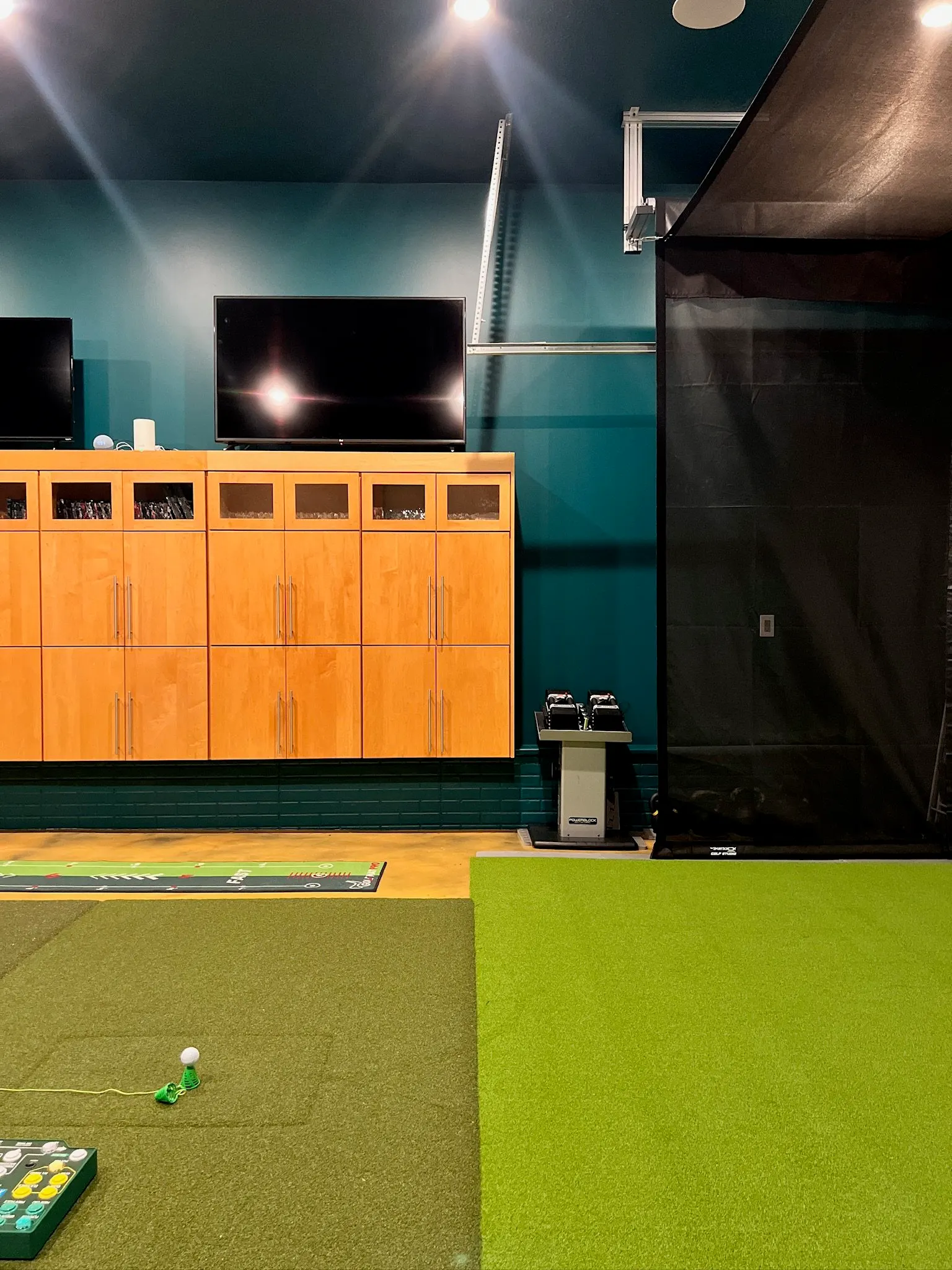 Indoor golf simulation room with a green mat, a golf ball and tee, wooden cabinets, two large mounted TVs, and gym equipment.