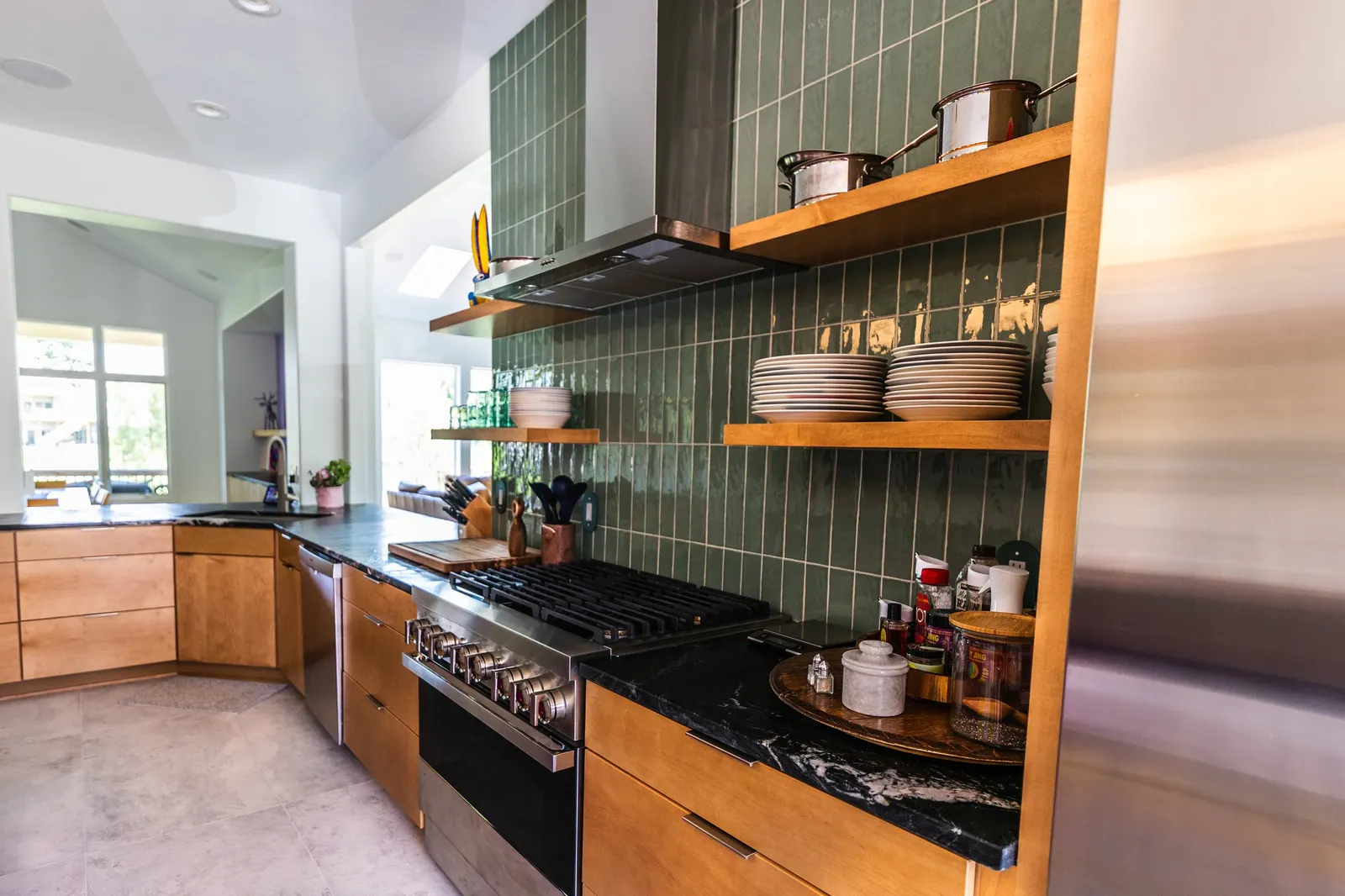 Modern kitchen with wooden cabinets, green tiled backsplash, stainless steel stove, open wooden shelves with bowls, pots, and glasses, and a black countertop with spices and kitchen tools.