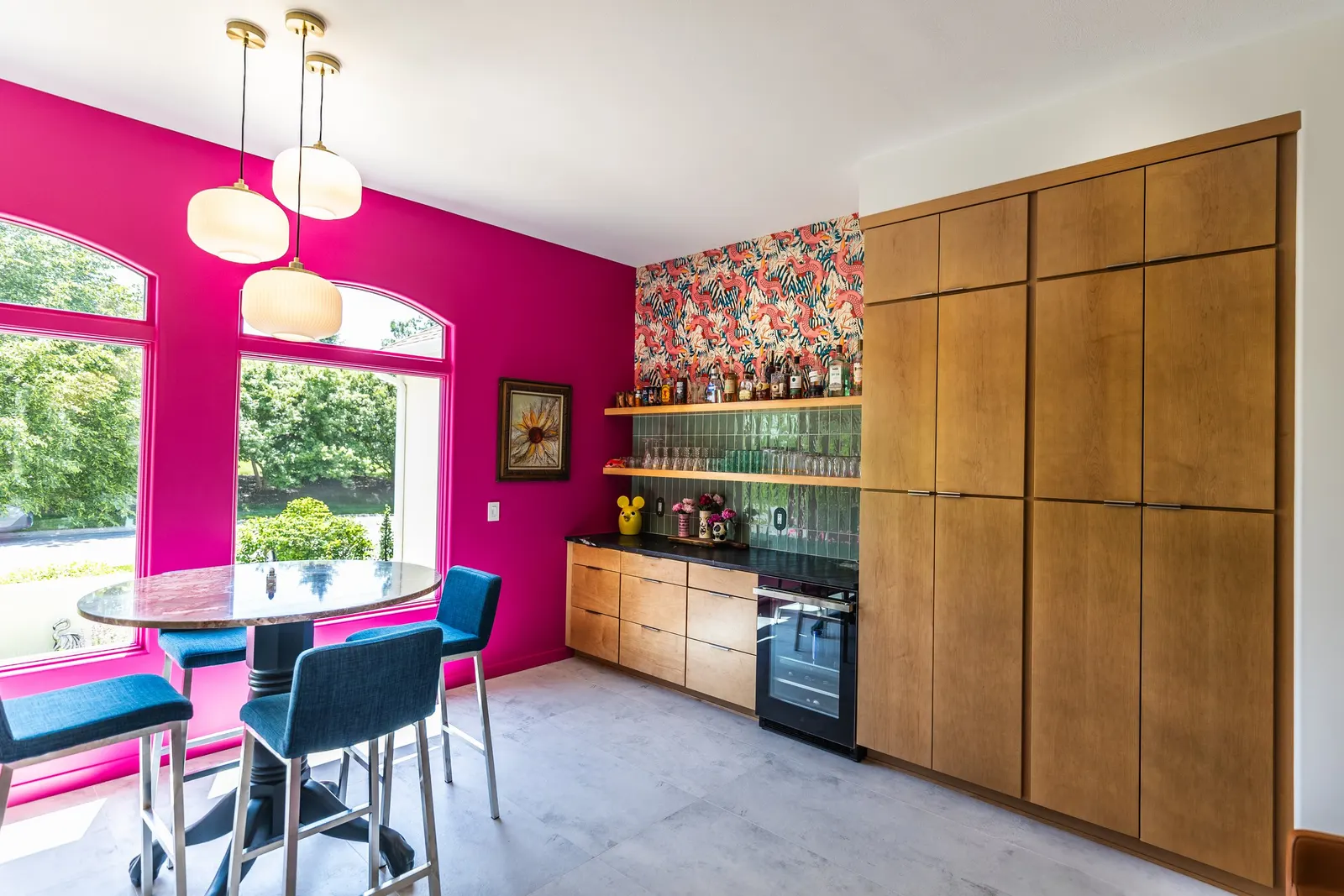Bright kitchen with vibrant pink wall, large windows, a ceiling light fixture with three white pendant lamps, a wooden table with three blue chairs, and a wooden cabinet with a black countertop, green tiled backsplash, and decorative items.
