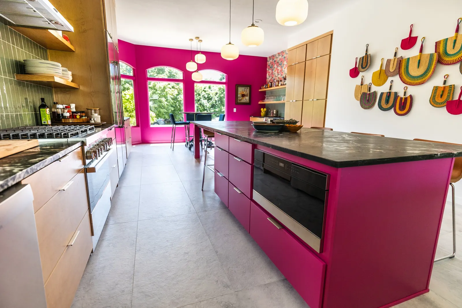 Modern kitchen with pink cabinetry and a large marble kitchen island, decorated with colorful woven bags on the wall, with bright pink walls and large windows showing green trees outside.