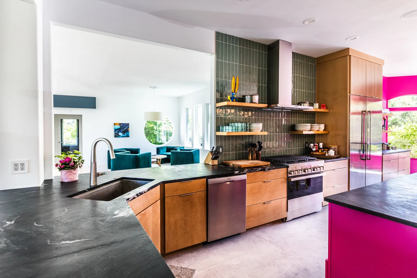 Modern kitchen with black marble countertops, wooden cabinets, green tiled backsplash, pink side wall, and a view into bright living room with blue chairs and large windows.