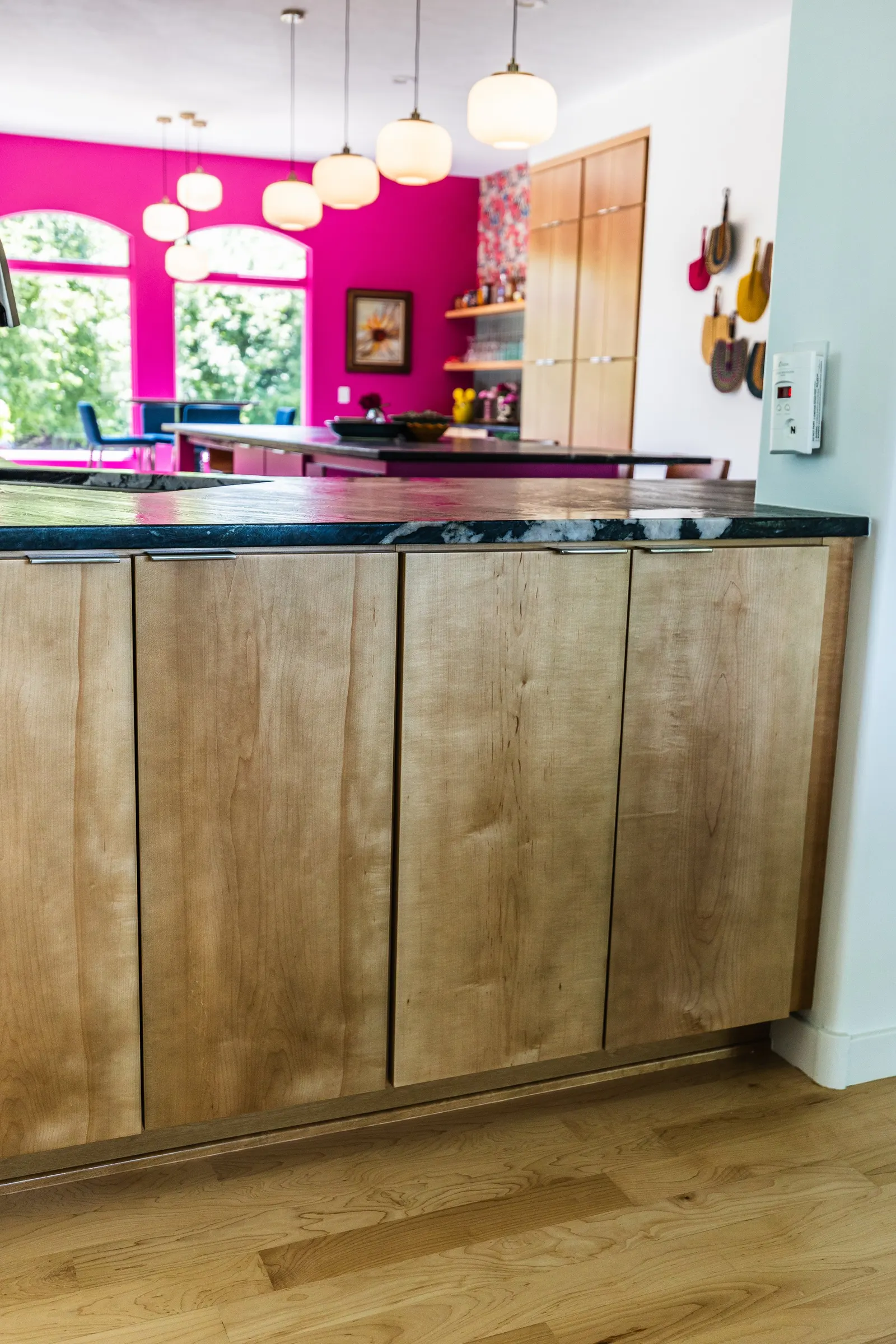 Close-up of a kitchen island with a wooden cabinet and black marble countertop in a modern kitchen with pink accent wall and hanging lights.
