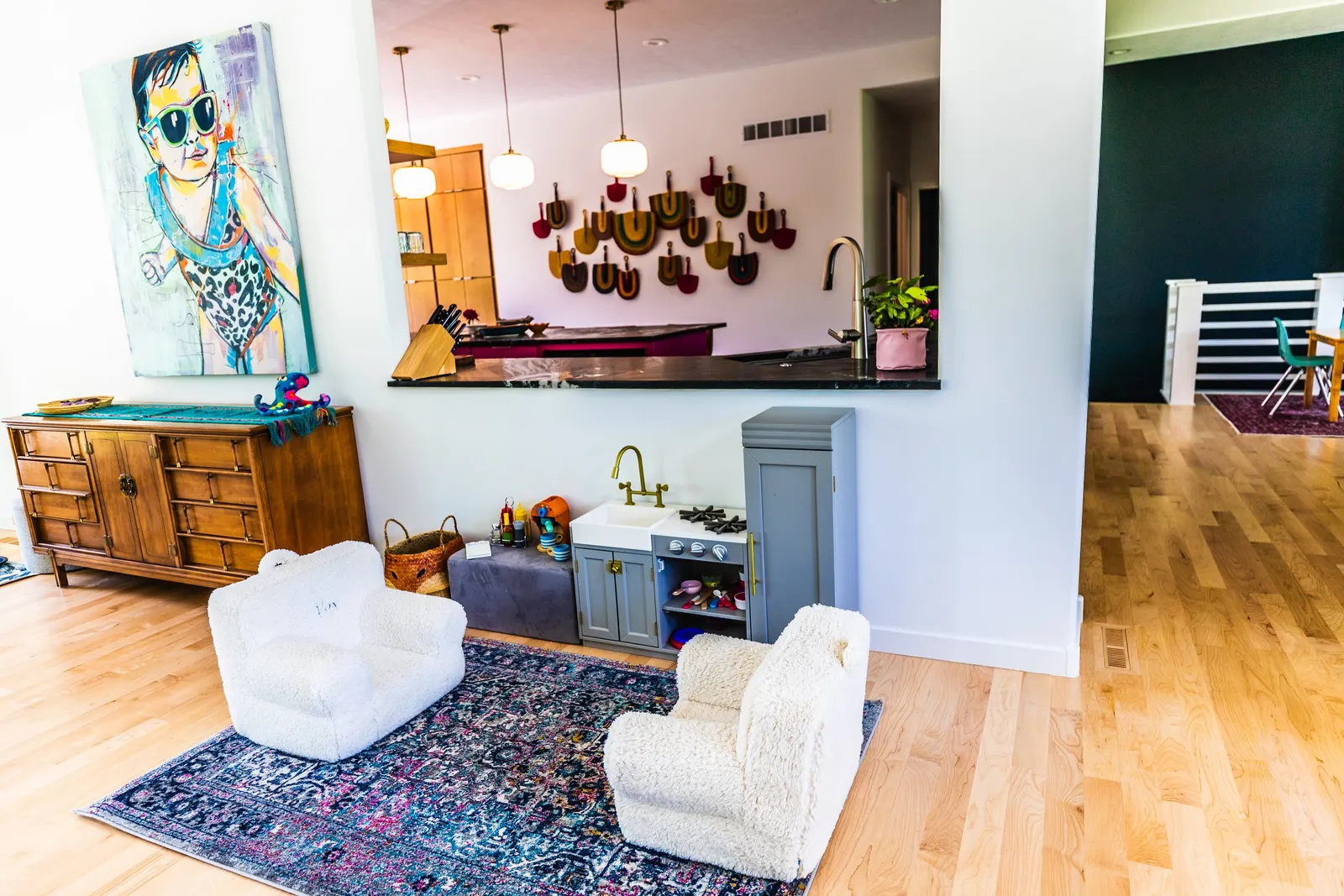 A children's play area with small chairs and a play kitchen on a colorful rug, with artwork, wooden furniture, and barstools visible in an open-concept living space.