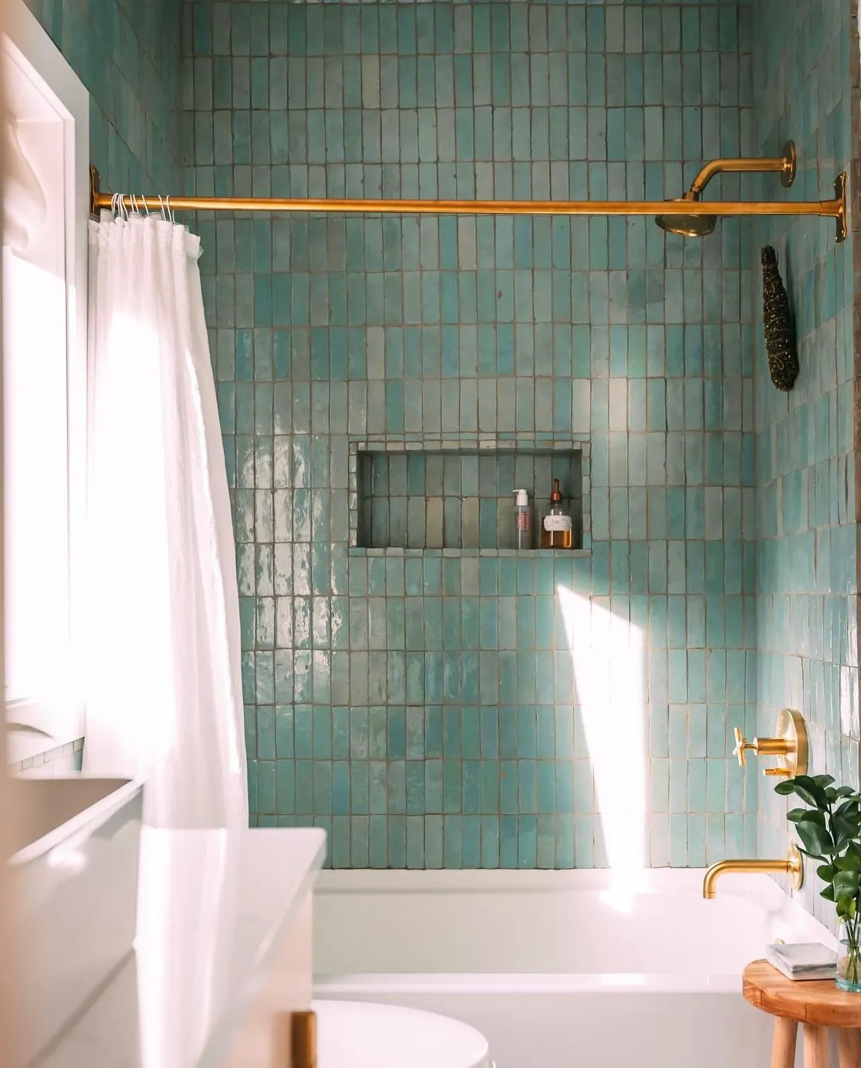 A bathroom with a tiled shower area featuring greenish-blue tiles, a gold shower rod, white curtain, built-in shower shelf with bottles, gold fixtures, and a small wooden table with a vase of green leaves.