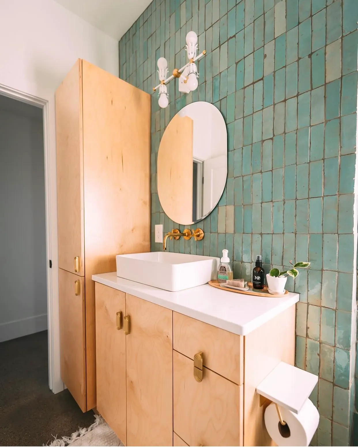Bathroom with a wooden vanity, white vessel sink, round mirror, green tiled wall, gold faucet and light fixture, small plant, soap dispenser, and toilet paper.