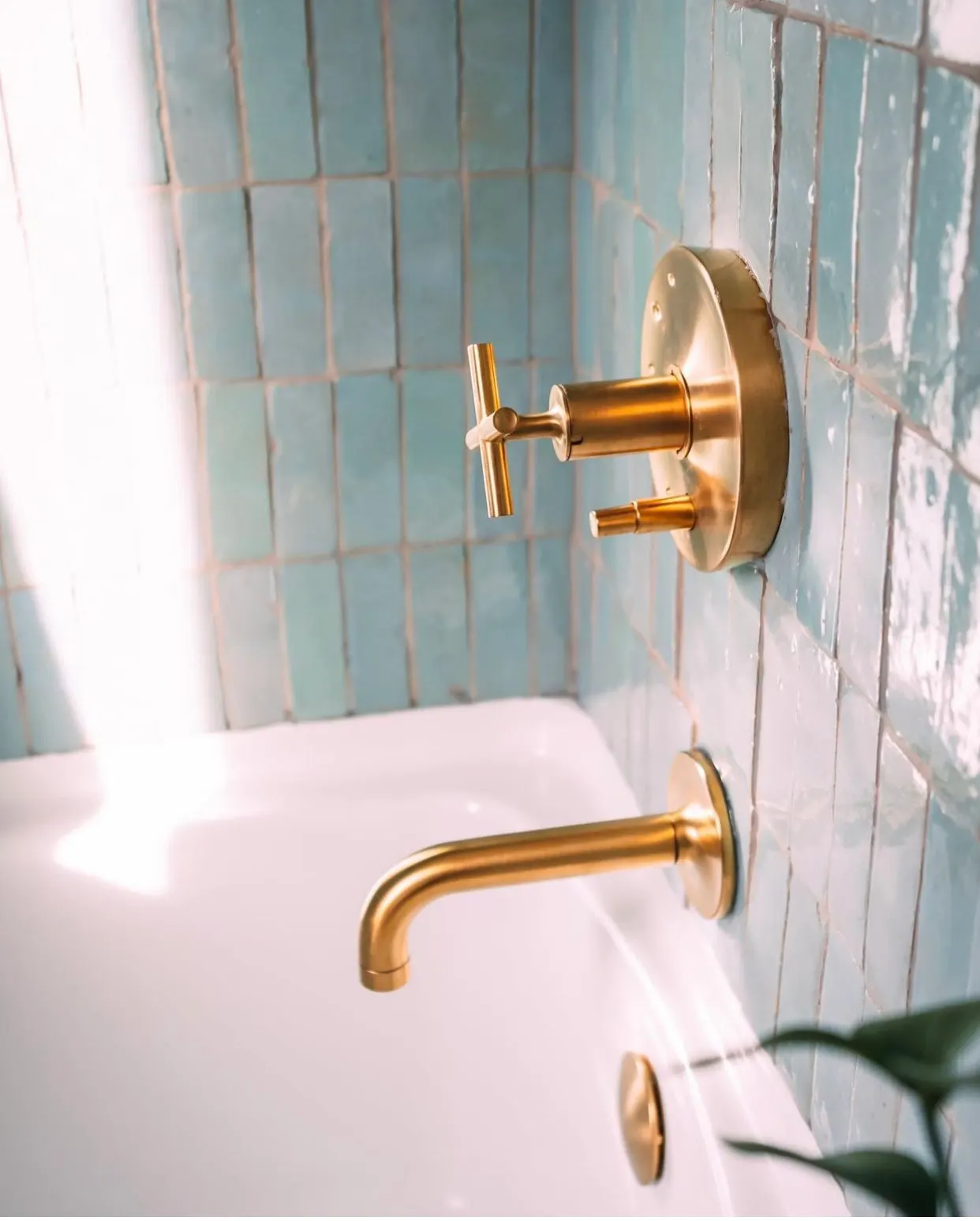 Close-up of gold-colored bathtub faucet, matching wall-mounted faucet handle, and wall-mounted soap dispenser, against a background of blue tiled bathroom wall.