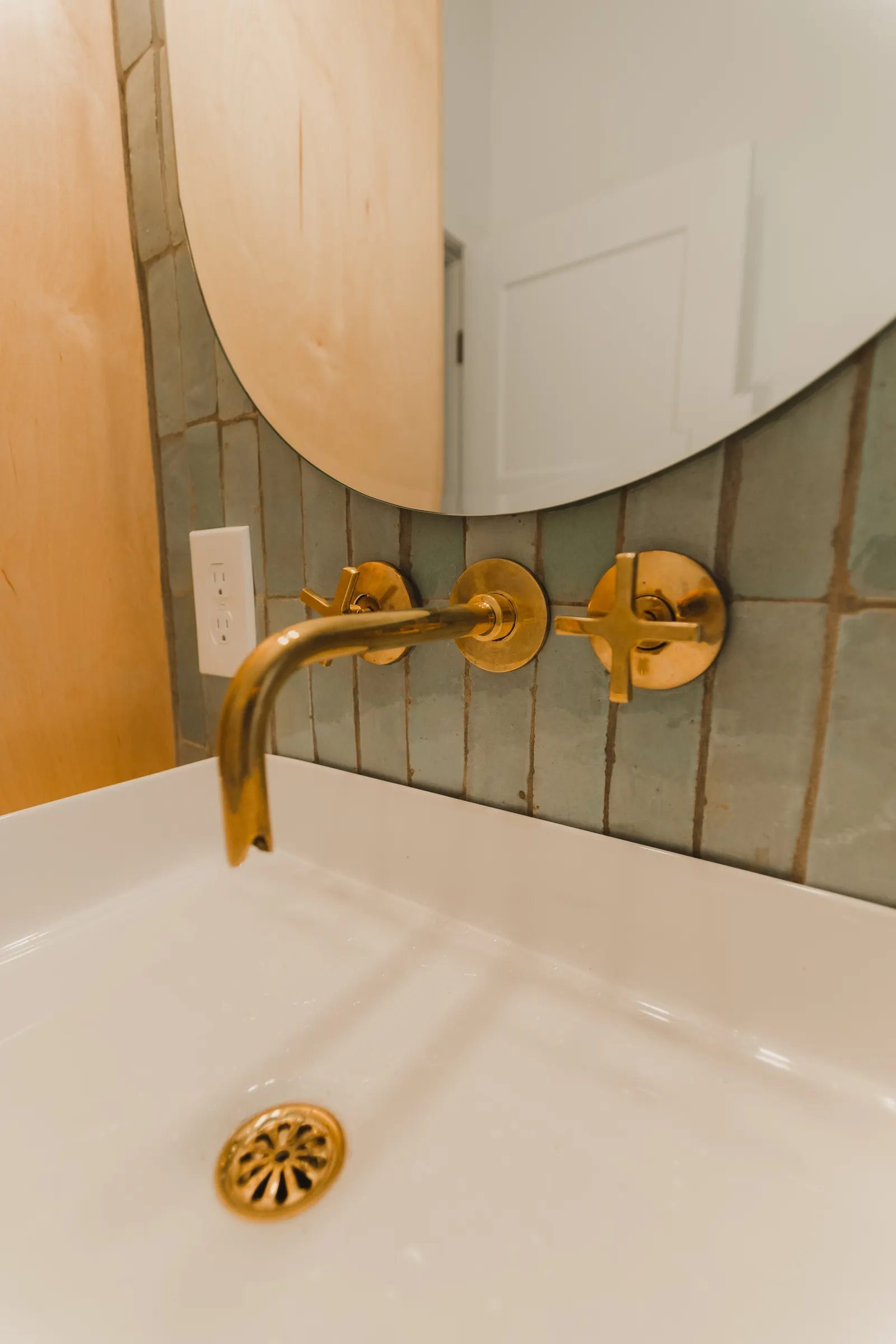 Bathroom sink with a gold faucet, a round mirror above, a black tile wall, and a white wall cabinet reflected in the mirror.