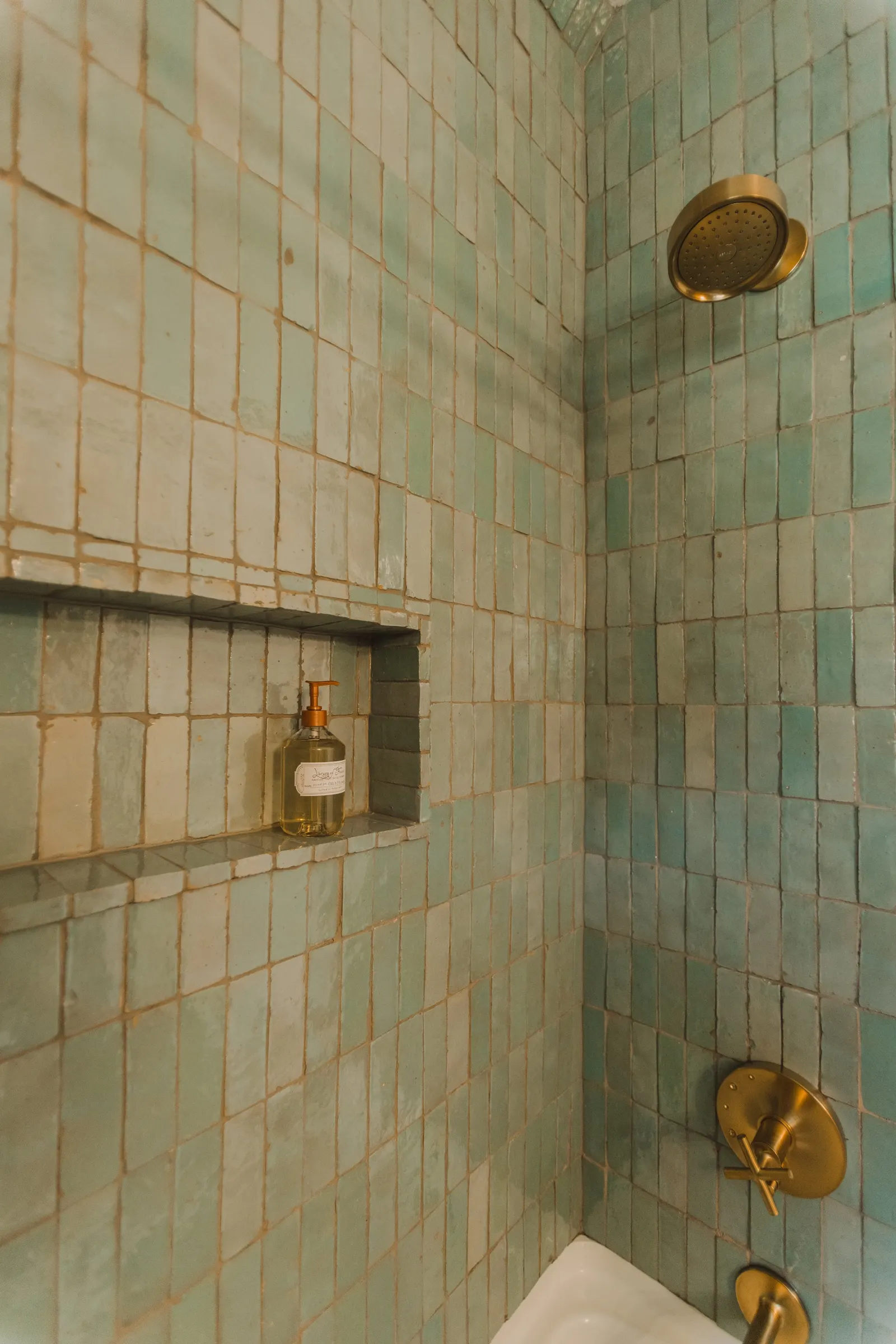 Inside a tiled shower with small green and beige tiles, a built-in shelf with a bottle of liquid soap, and brass fixtures including a showerhead and a control handle.