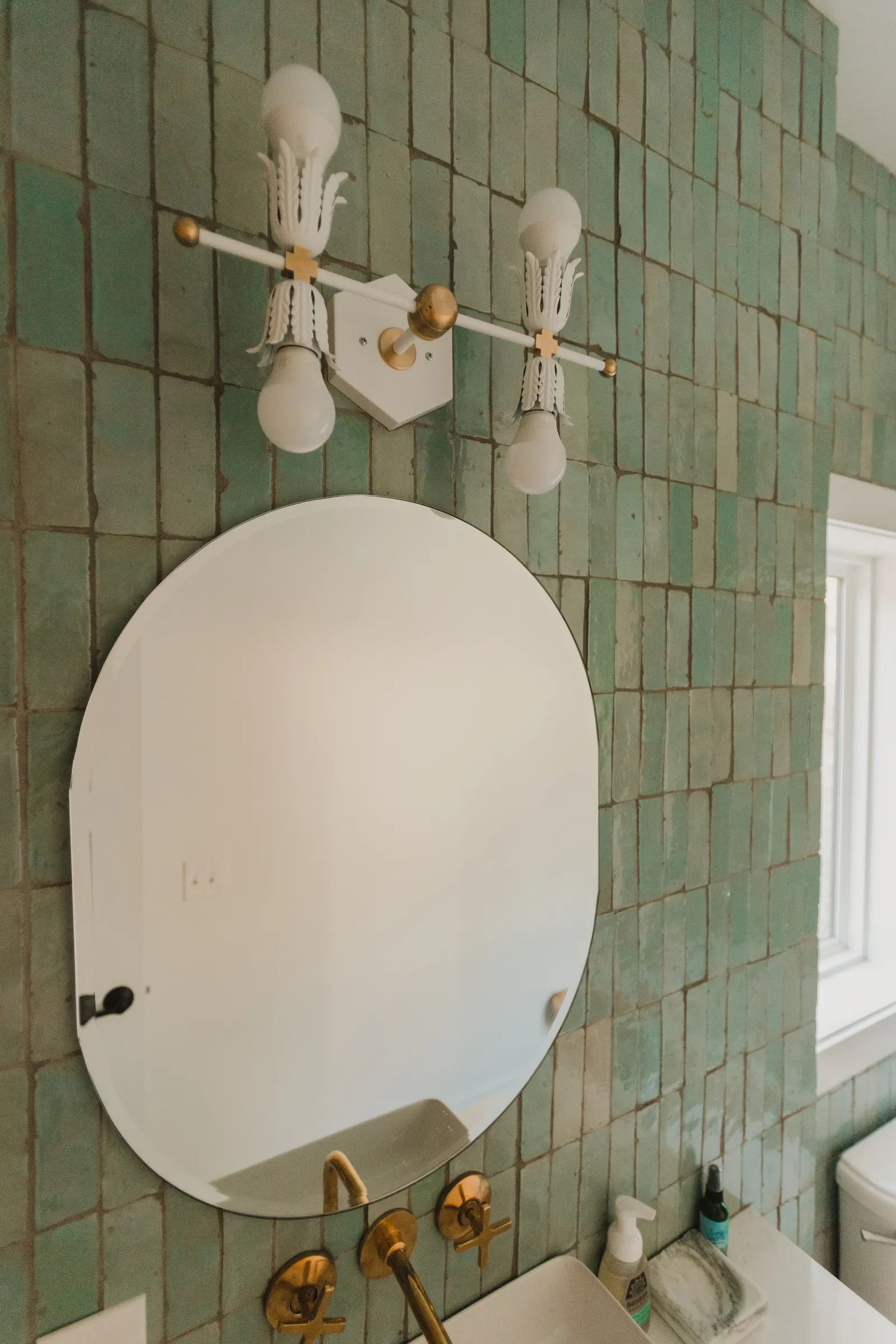 Bathroom wall with green tiles, a round mirror, a brass faucet, and a light fixture with three bulbs.