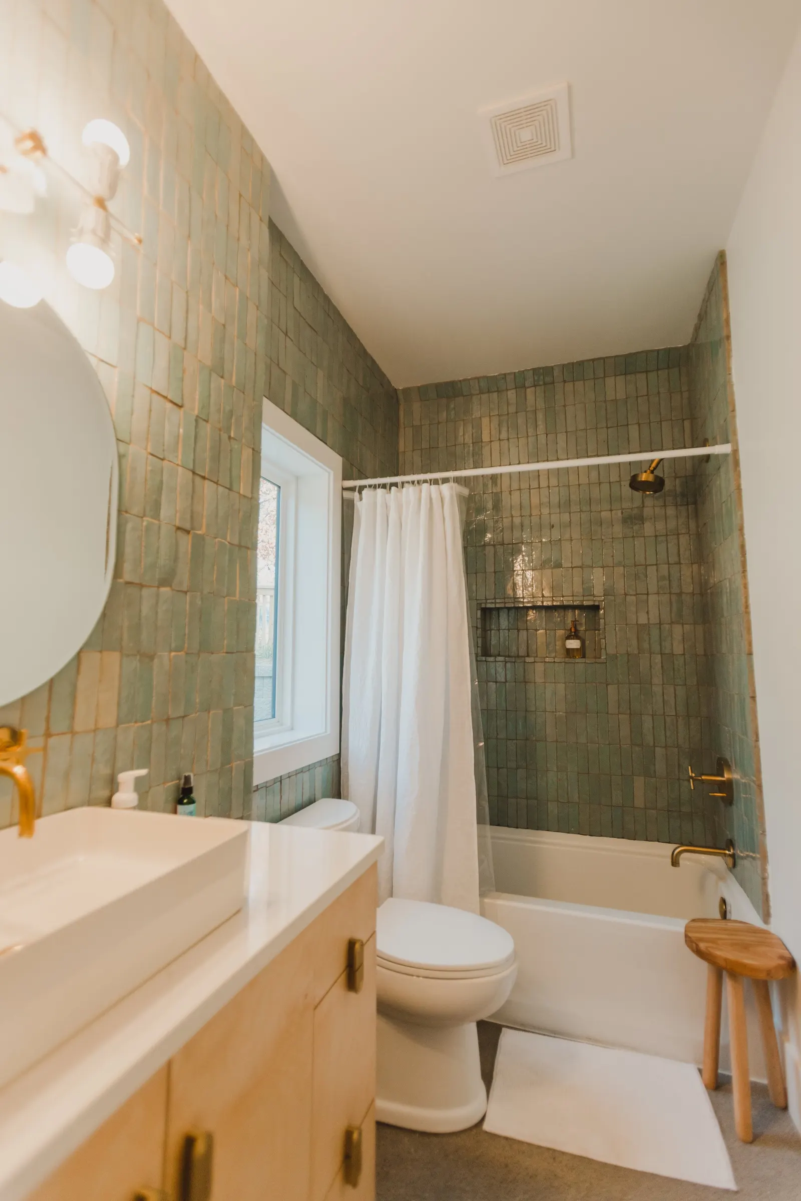 Bathroom with green tiled walls, a white bathtub with a shower, a window, a white curtain, a toilet, a vanity with a vessel sink, a mirror, and a small wooden stool.