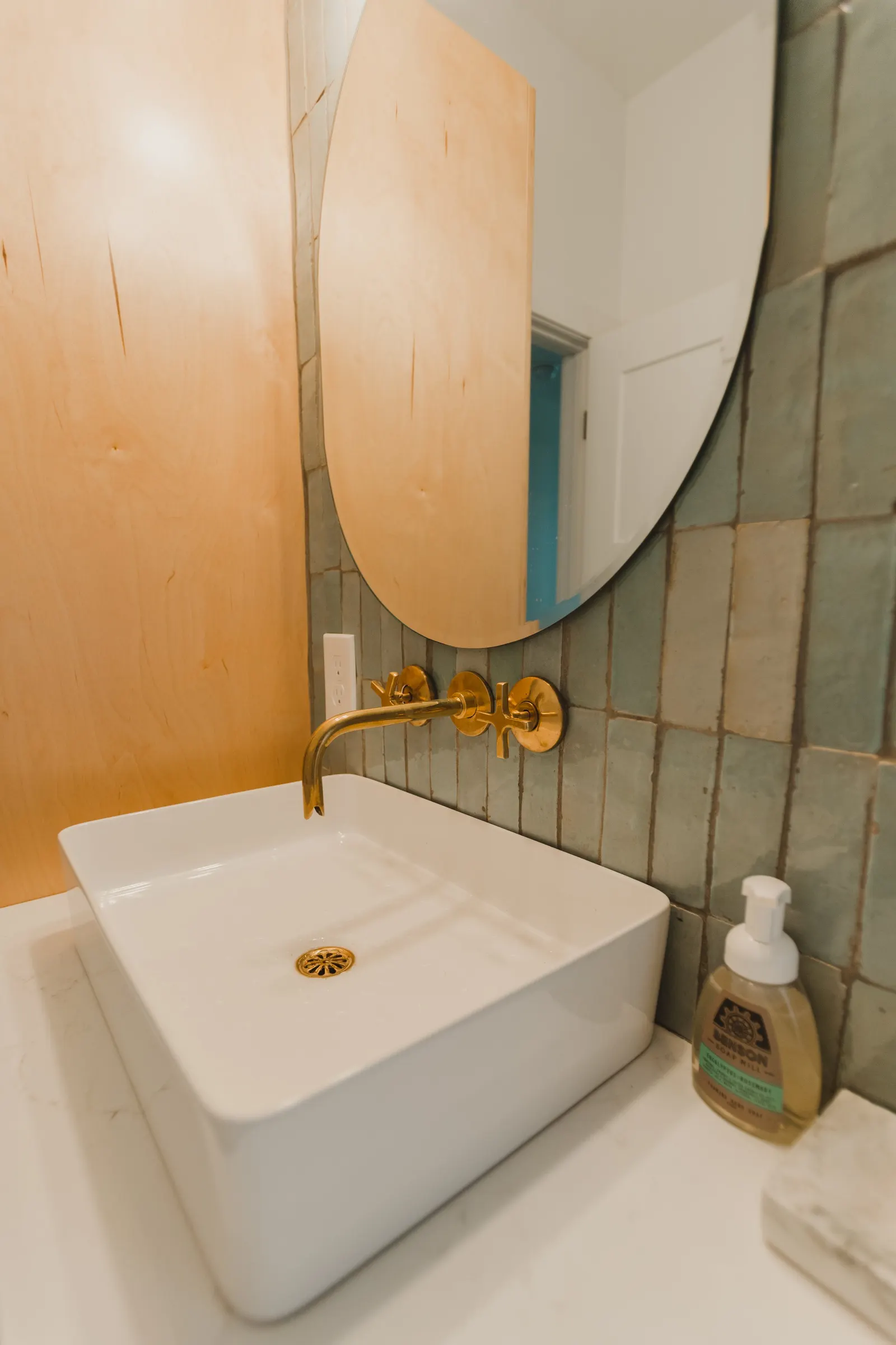 A bathroom sink with a rectangular white vessel, brass faucet and fixtures, a round mirror, a soap dispenser, and a wooden wall with green tiles.