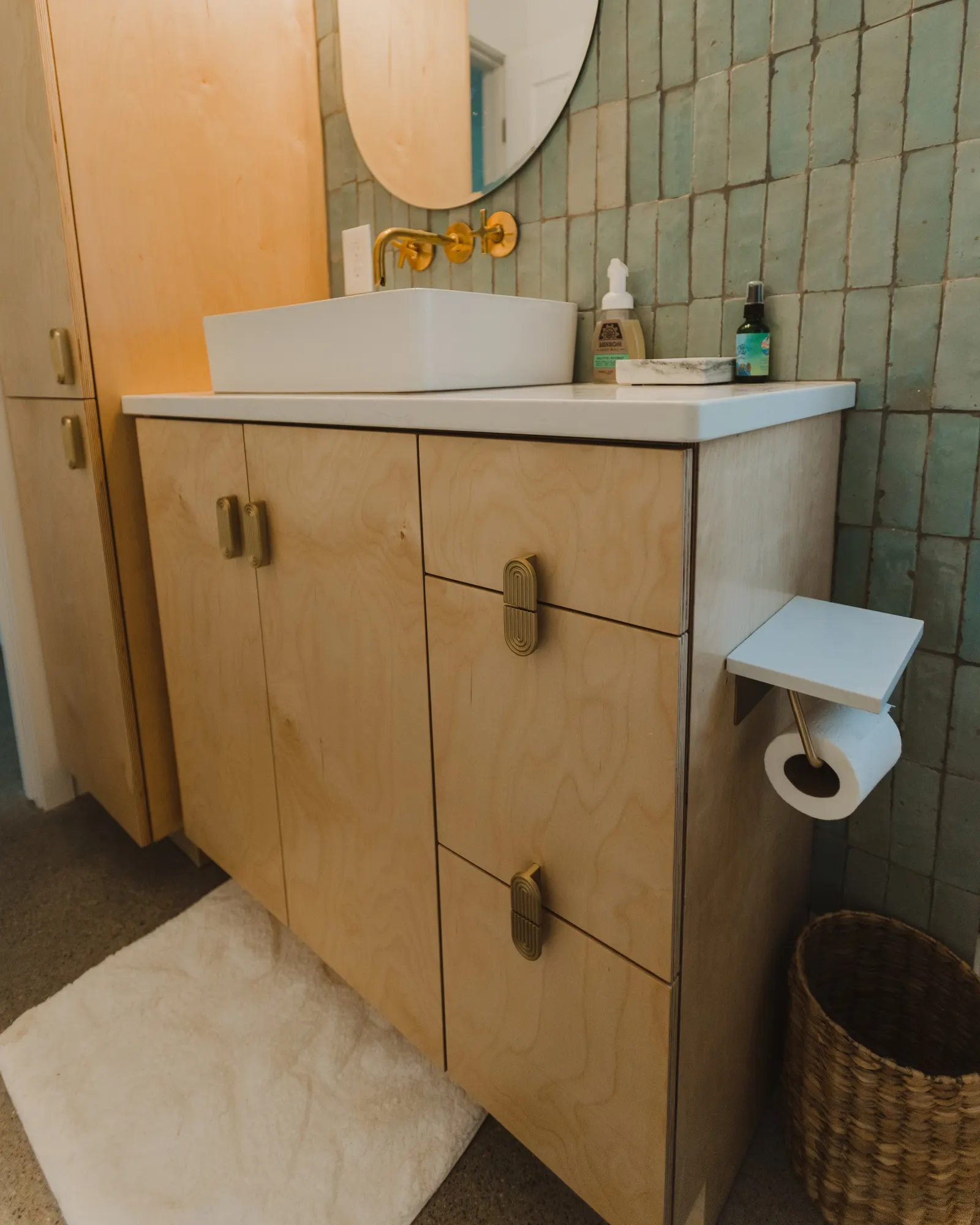 Wooden bathroom vanity with a white rectangular sink, gold faucet, mirror, soap dispenser, sanitizer, and spray bottle. A white toilet paper roll holder attached to the side, a wicker basket, and a white bath mat on the floor. Green tiled wall behind