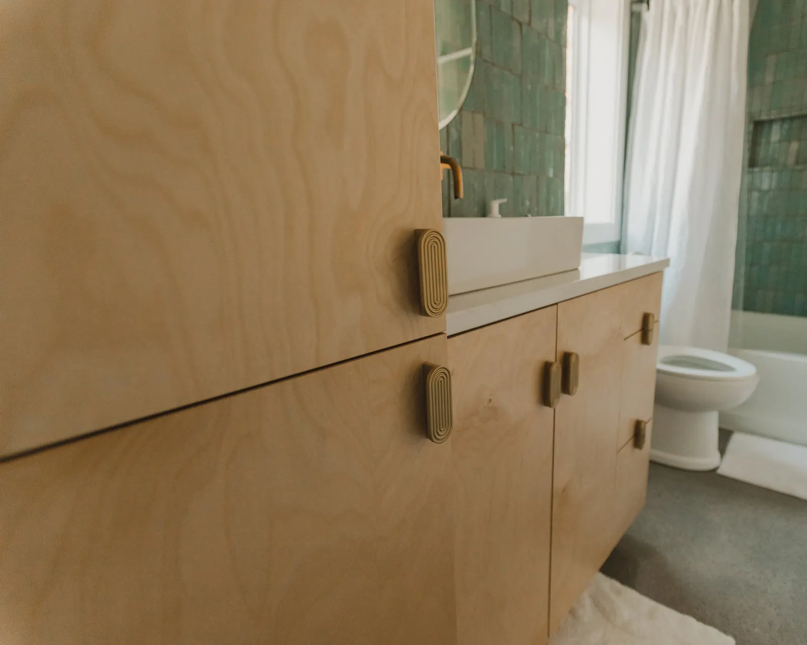 Close-up of a light wooden bathroom cabinet with rounded handles, next to a white sink, in a bathroom with green tiled walls, a window with white curtains, a toilet, and a bathtub with a shower curtain.