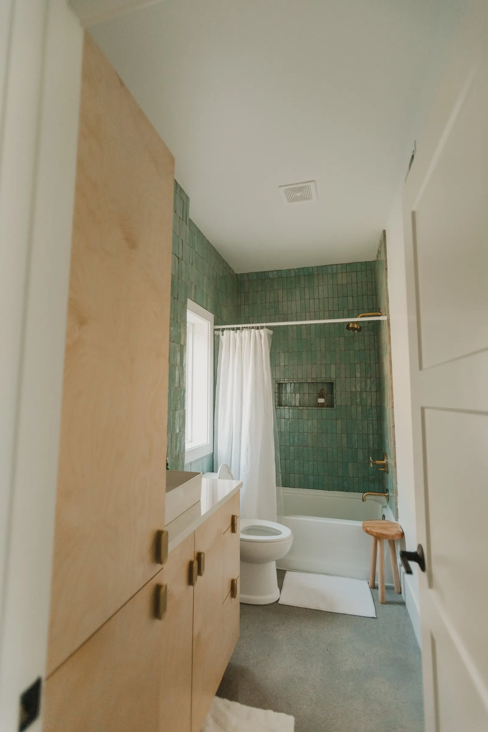 View of a bathroom seen through an open door, featuring a wooden vanity with a sink, a toilet, a bathtub with a green tiled wall, a window, a shower curtain, and a small wooden stool.